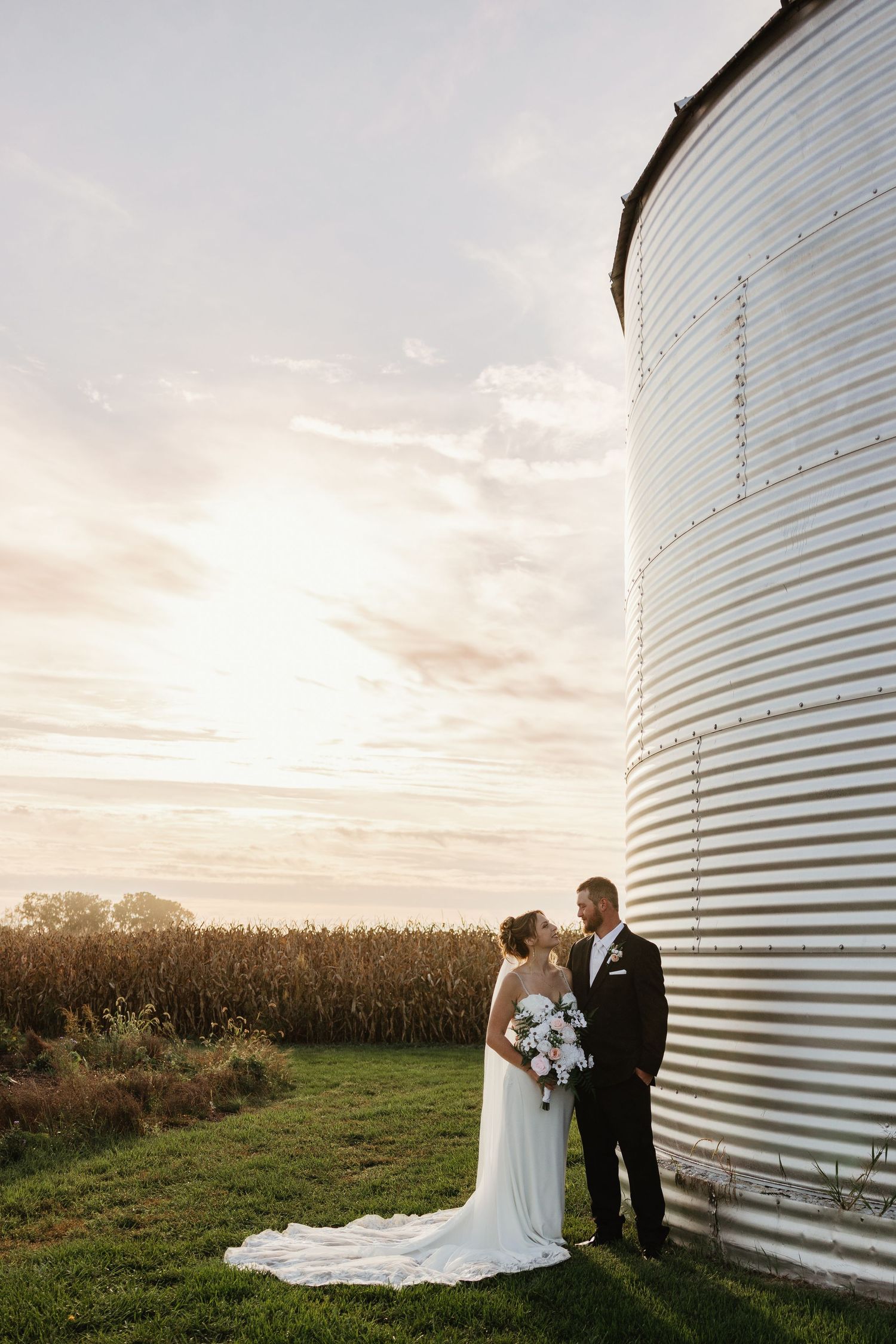 Bride and groom stand by a grain silo at sunset with a cornfield backdrop on their wedding day.