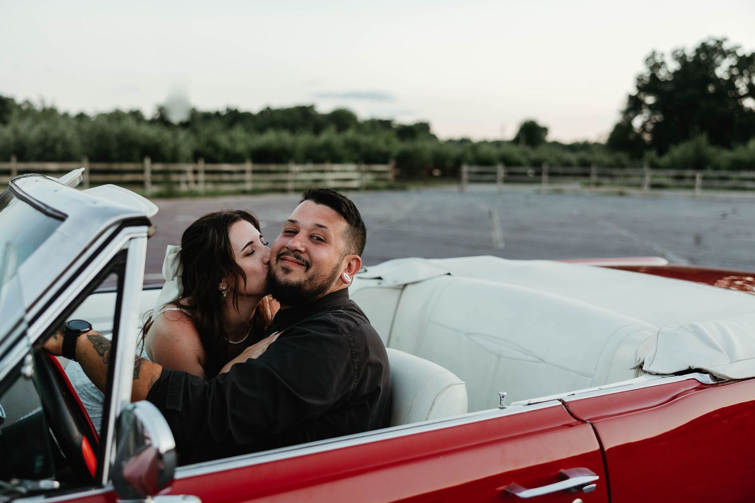 Couple shares romantic kiss in classic red convertible car at outdoor venue during golden hour.