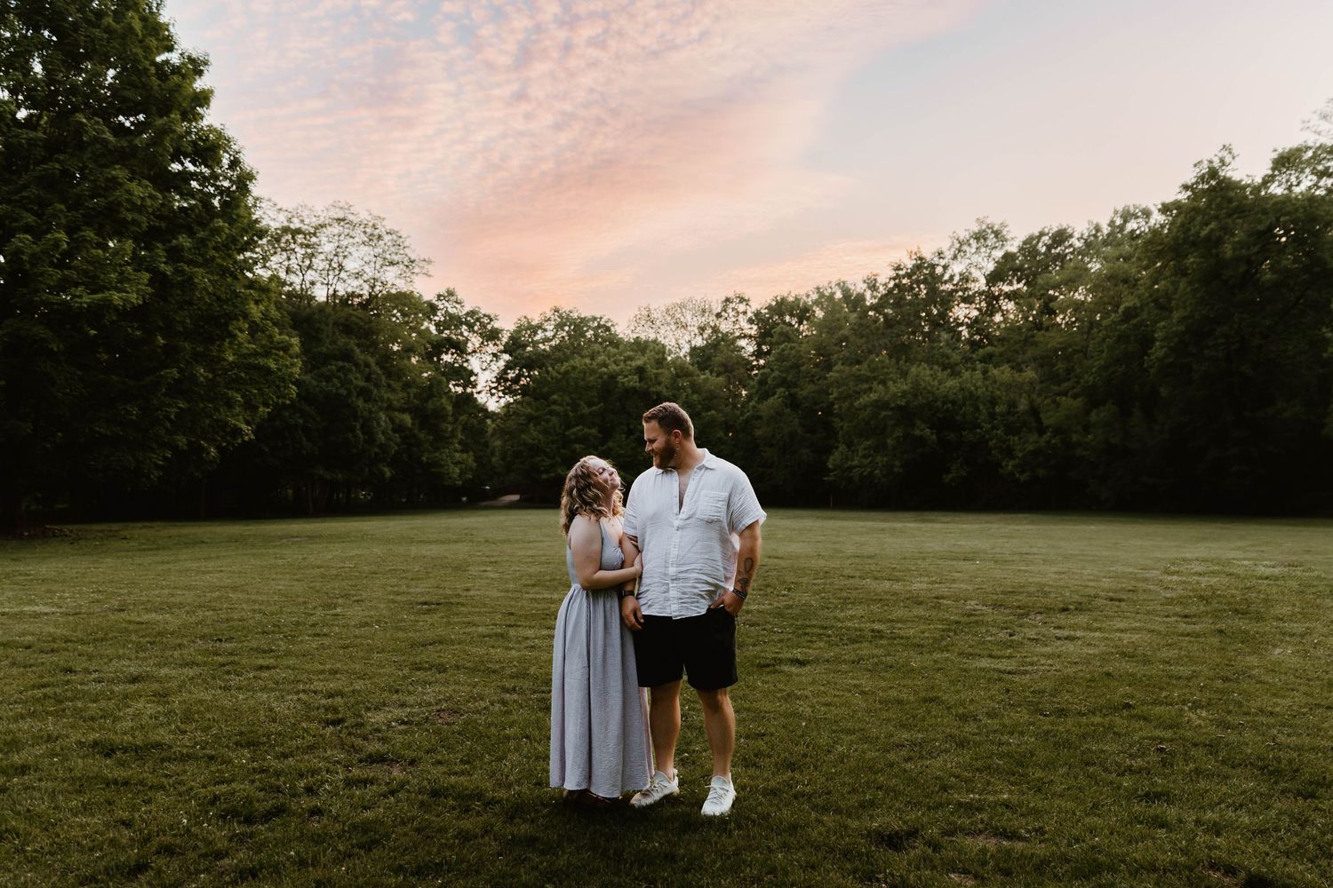 Couple standing together in a lush green field at sunset with pink sky and trees in background.