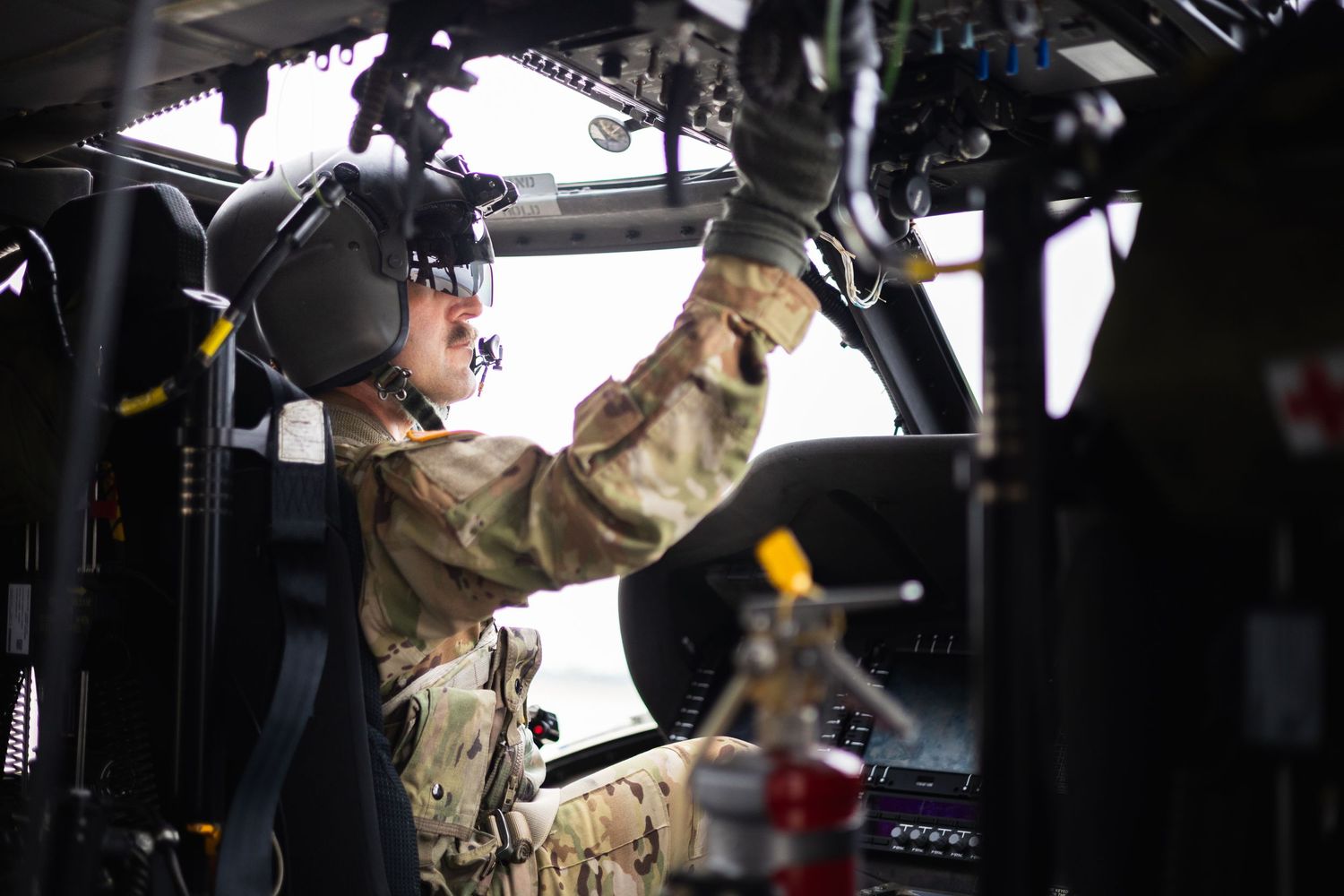 Military pilot in camouflage uniform performing pre-flight checks inside a helicopter cockpit.