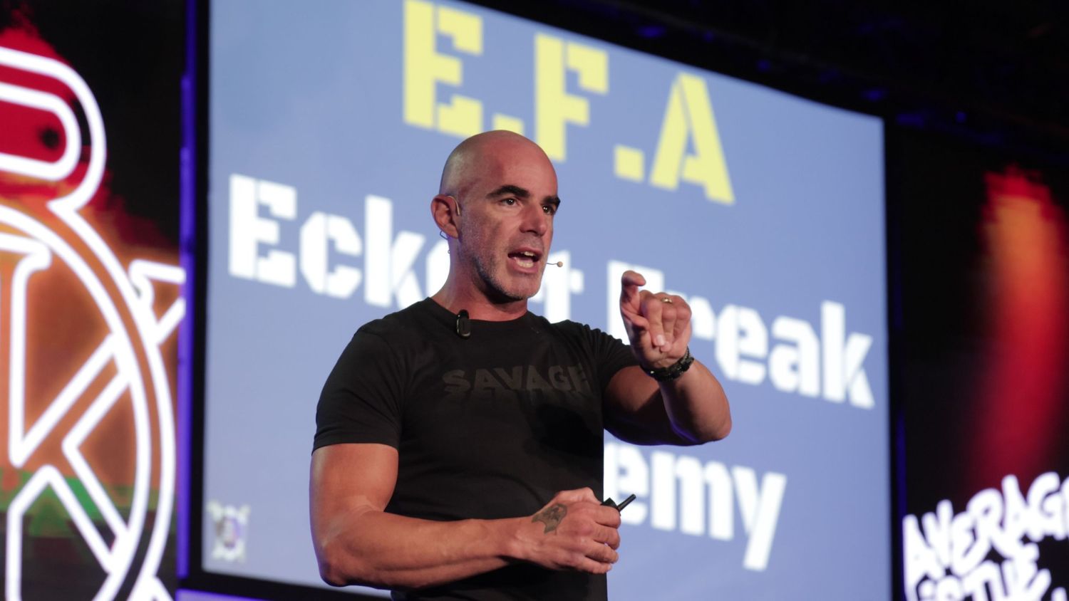 Bald man in black Savage shirt presenting at EFA Eckoff Break Academy event on stage.