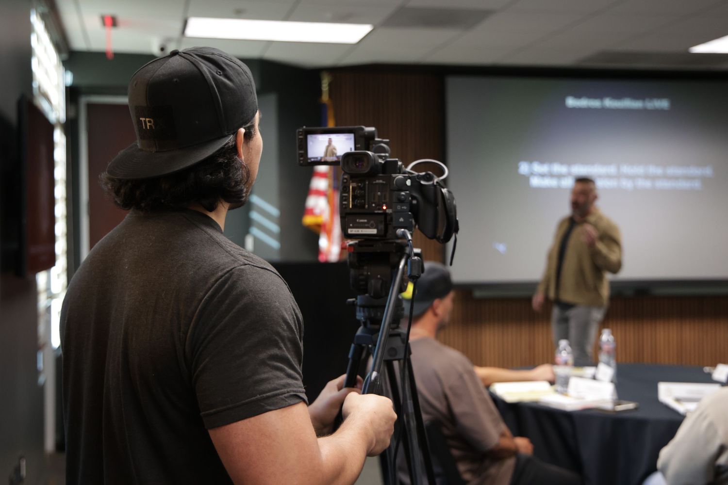Videographer filming speaker at professional seminar with camera on tripod in conference room.