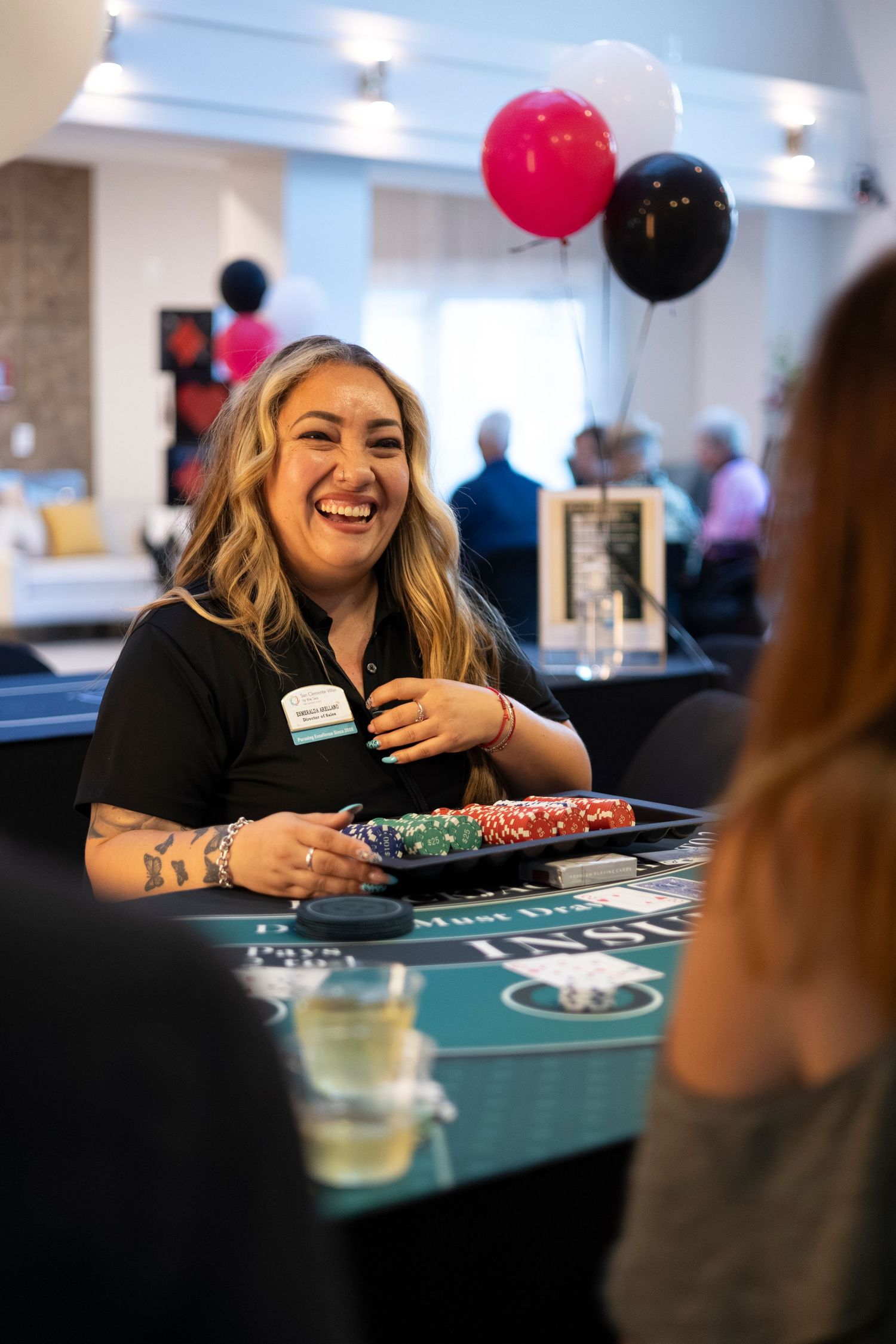 Smiling woman dealer with tattoos holding casino chips at blackjack table at party event.