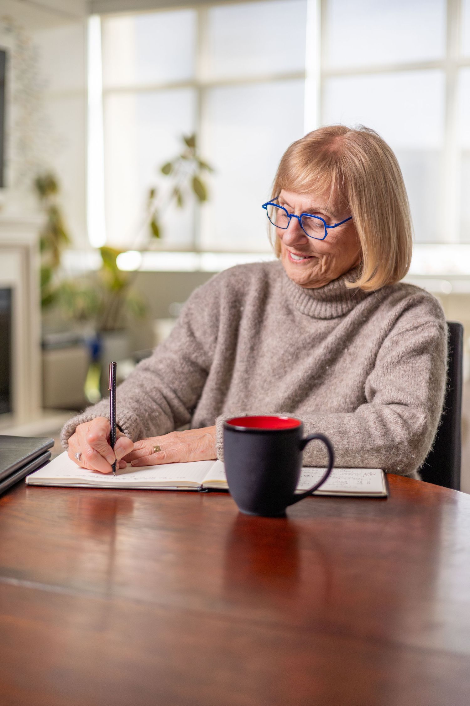 Senior woman writing in notebook at desk with coffee mug.