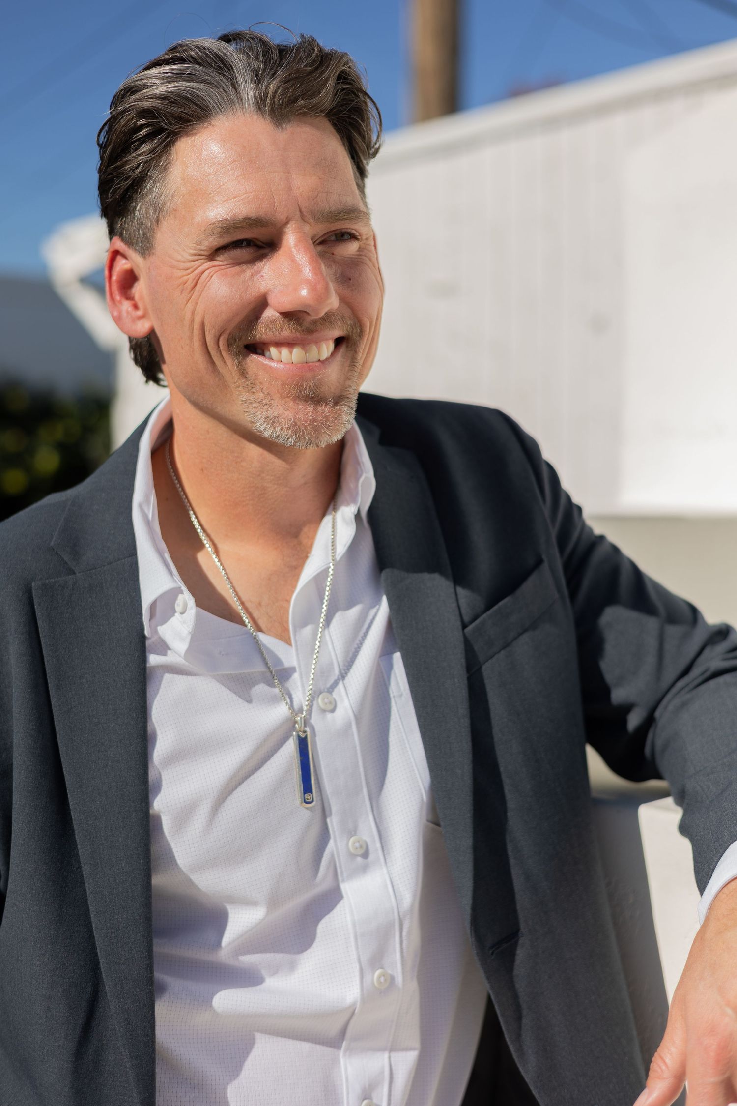 Smiling man in grey blazer and white shirt outdoors on sunny day.
