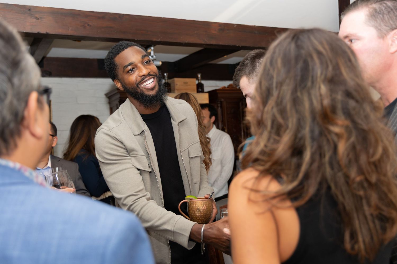 Tall Black man in beige jacket smiling and socializing at casual indoor networking event.