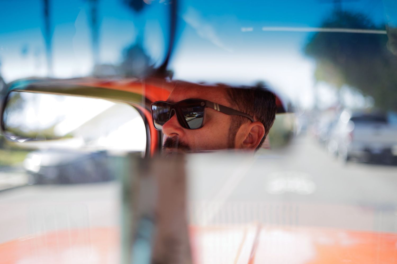 Man with sunglasses reflected in vintage car rearview mirror on sunny day.