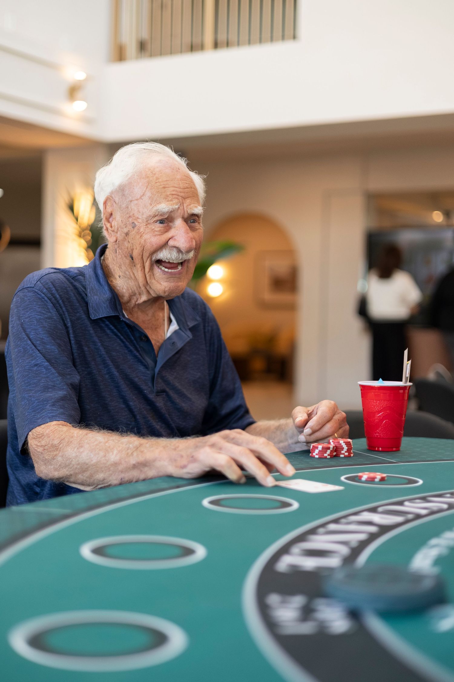 Happy elderly man laughing at casino blackjack table with poker chips and red cup.