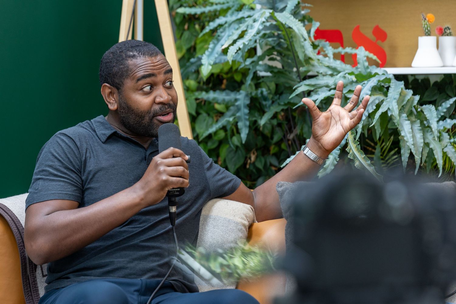 Man speaking into microphone during interview in green plant-filled studio.