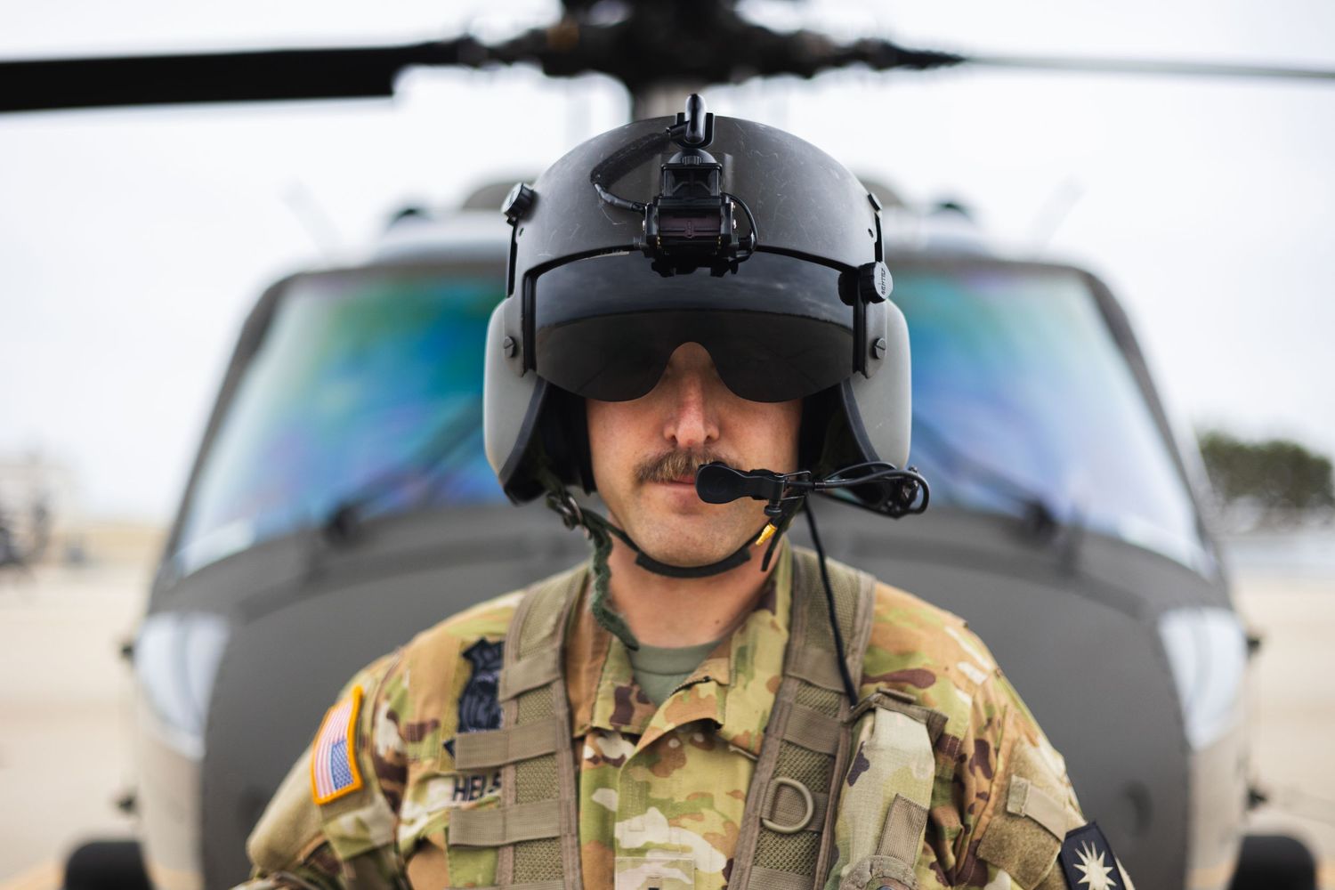 Military soldier in combat helmet standing in front of Black Hawk helicopter on airfield.