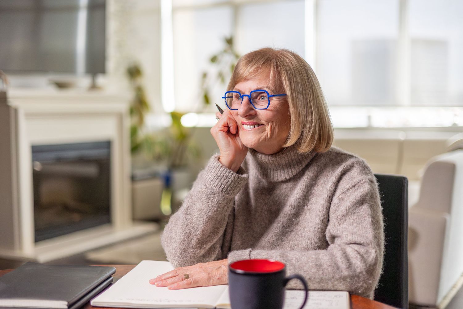 Smiling senior woman with blue glasses sitting at desk with red mug in bright living room.