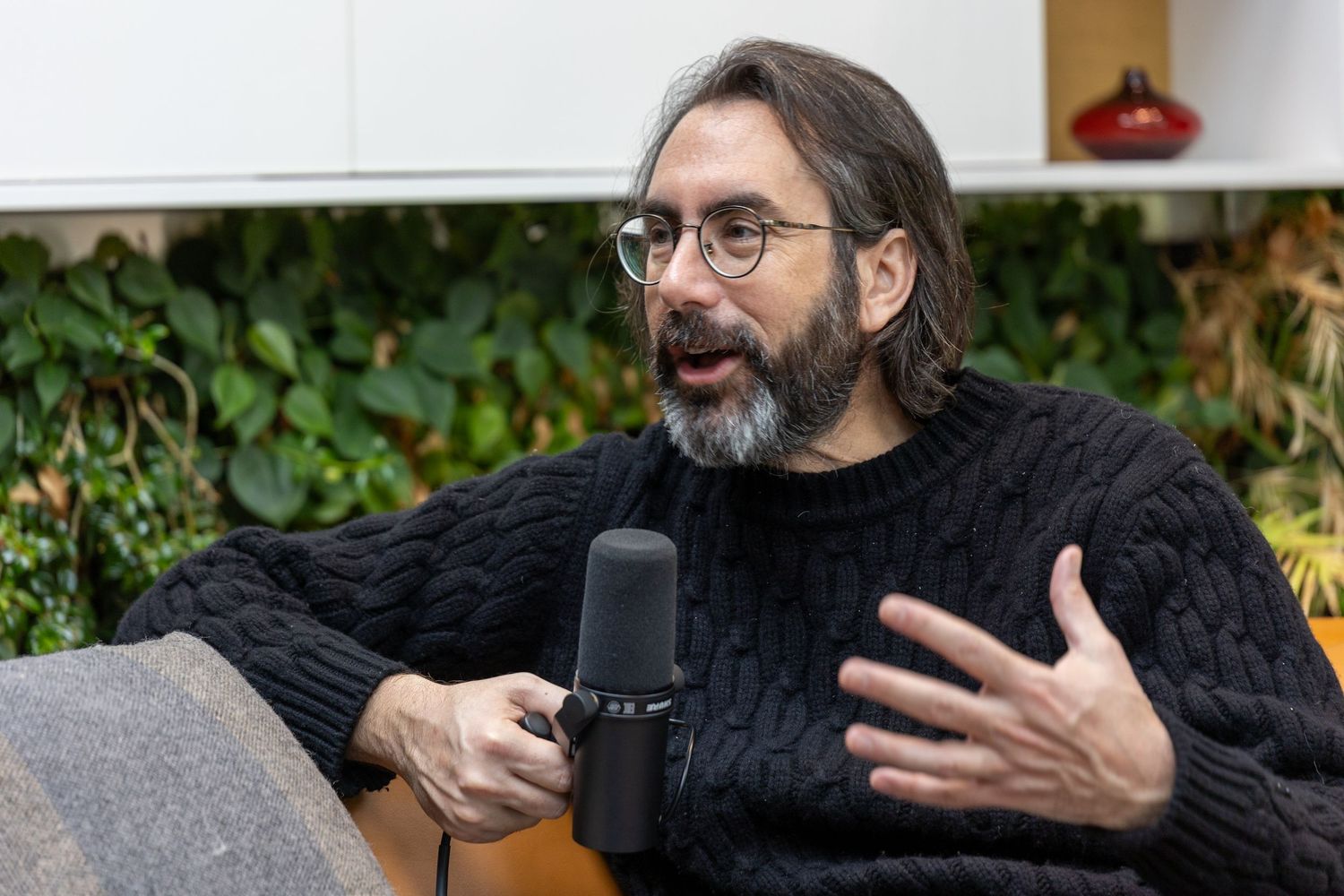 Bearded man with glasses speaking into podcast microphone indoors.