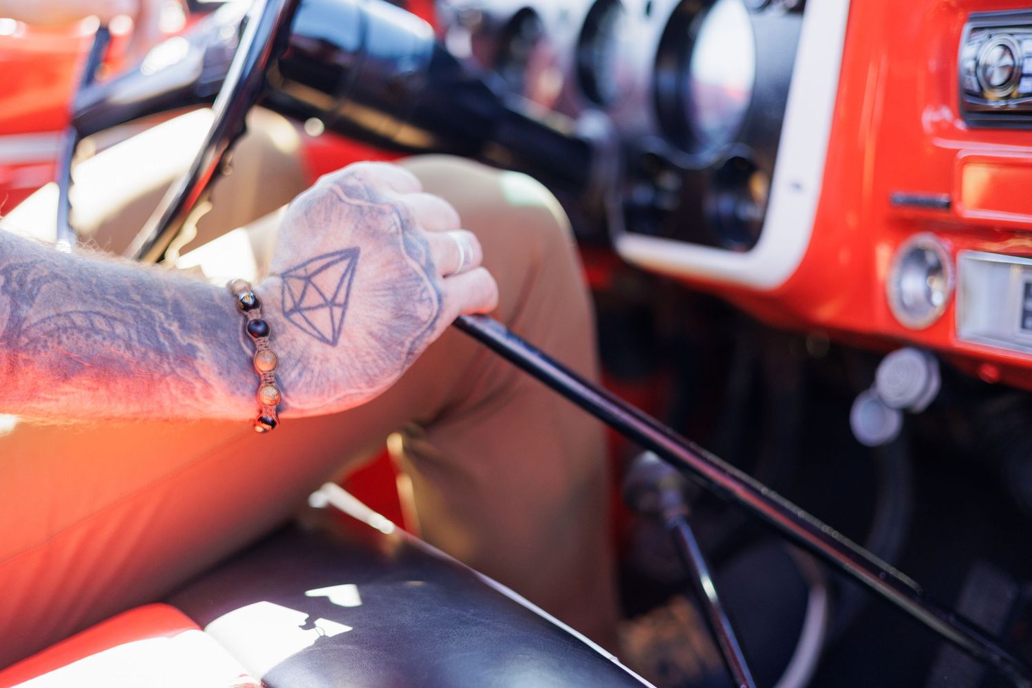 Tattooed hand with bracelet gripping a gear shift inside a vintage red car interior.