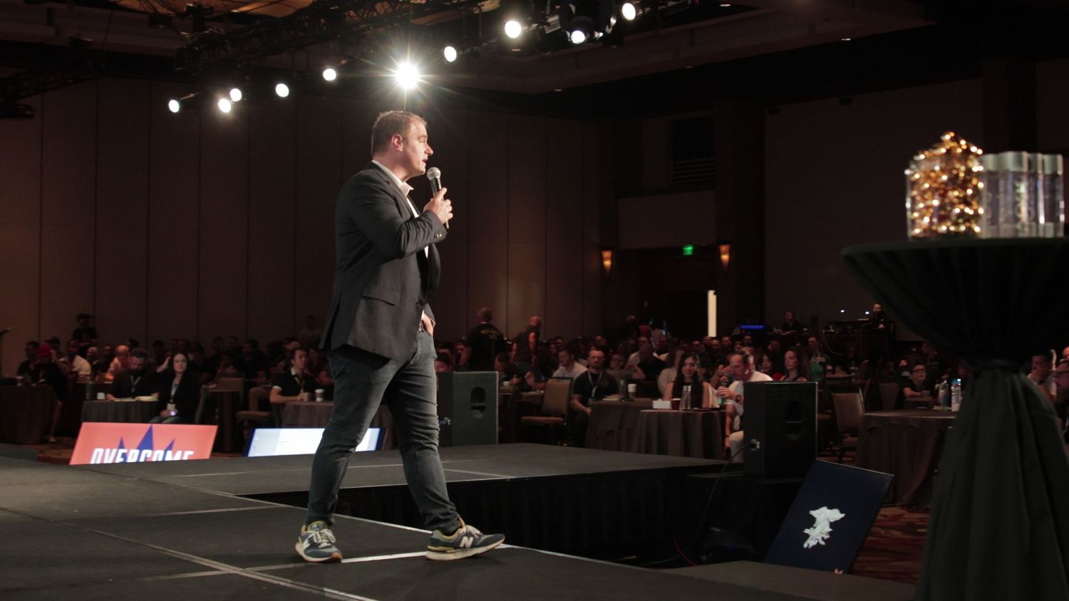 Male speaker in blazer presenting on stage to large audience at evening conference event.