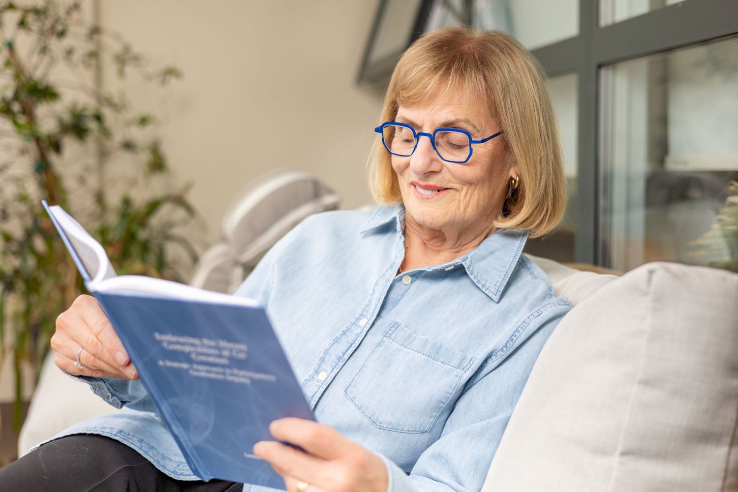Senior woman with blue glasses reading book on sofa at home.