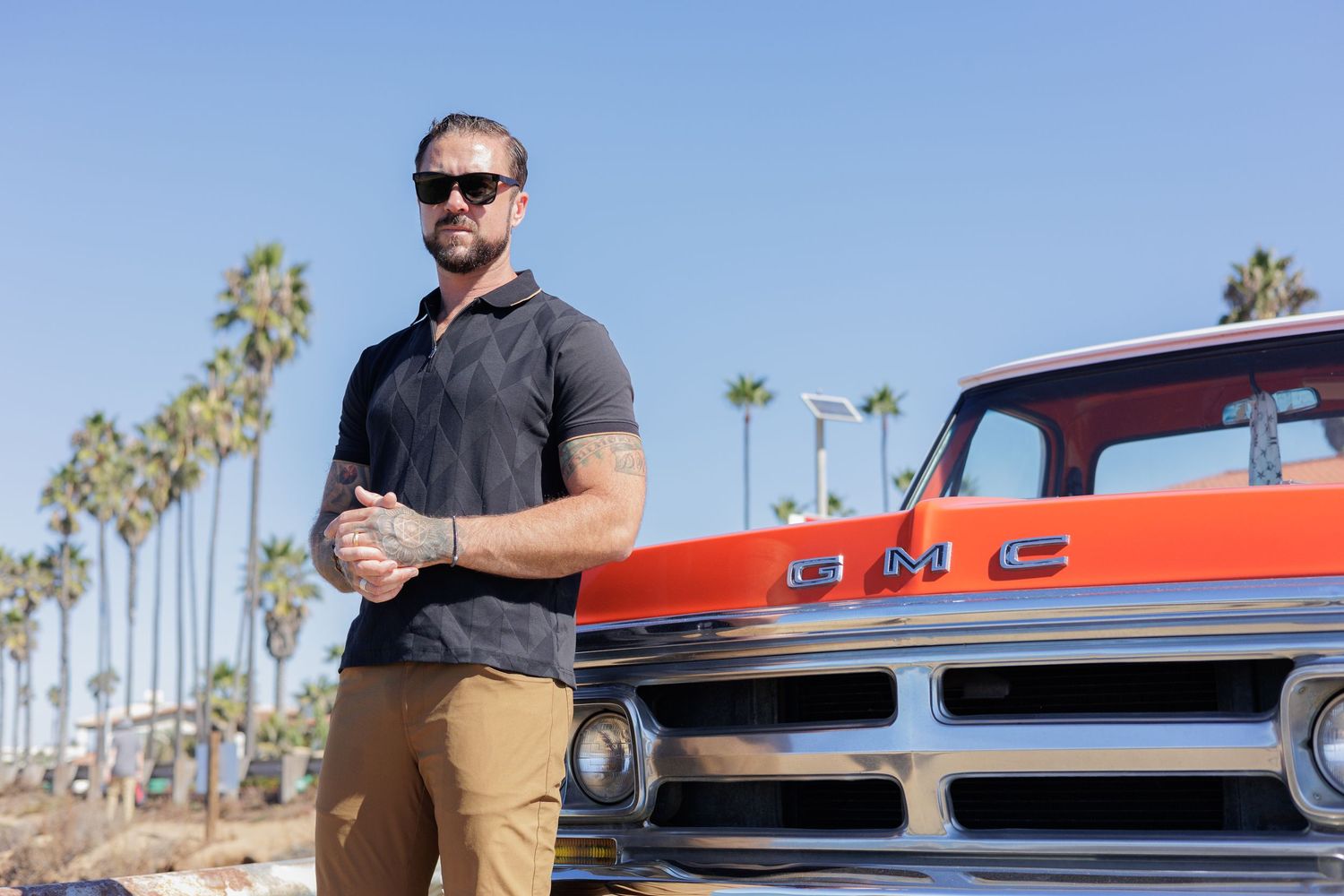 Man in sunglasses stands beside classic GMC truck with palm trees under blue sky.