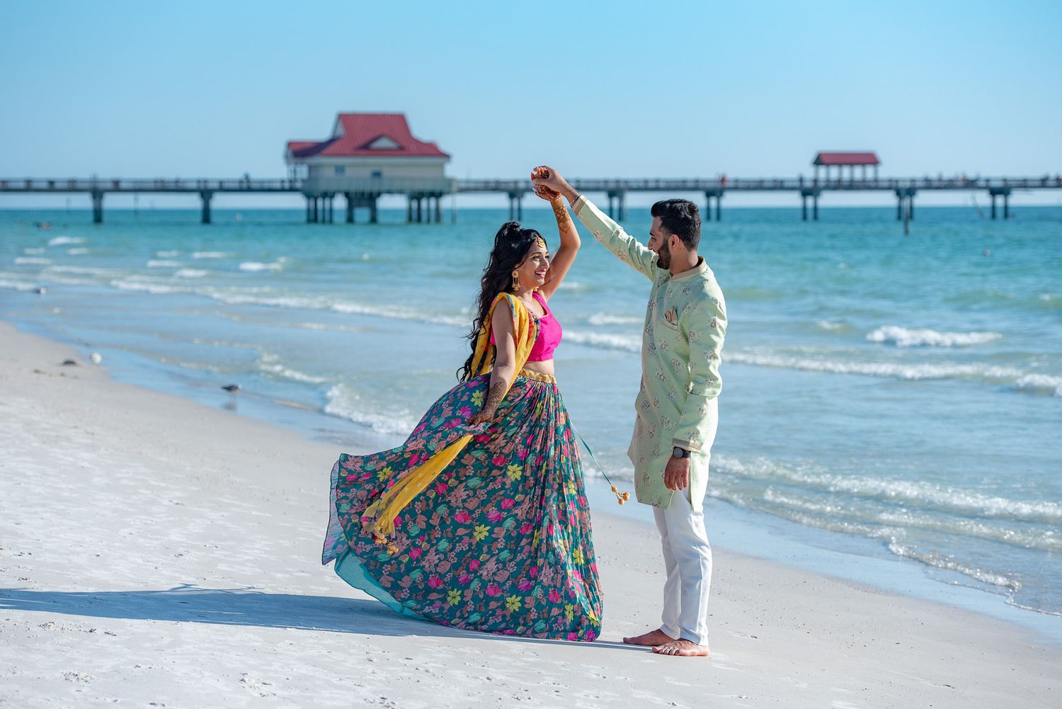 Couple dancing on beach with pier and red-roofed structure in background.
