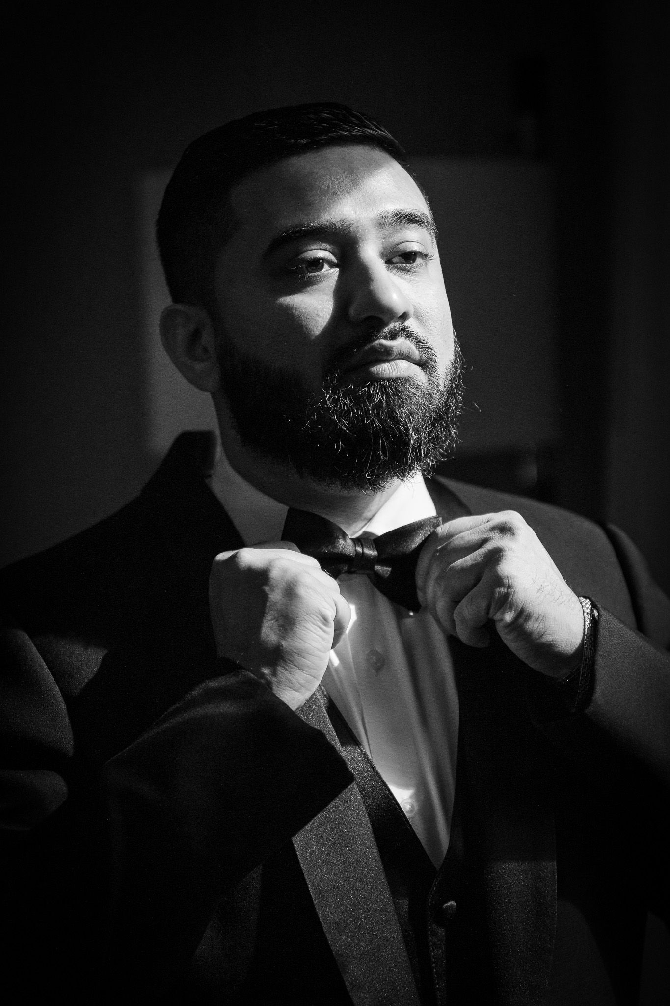 Black and white portrait of groom adjusting bowtie in dramatic lighting.