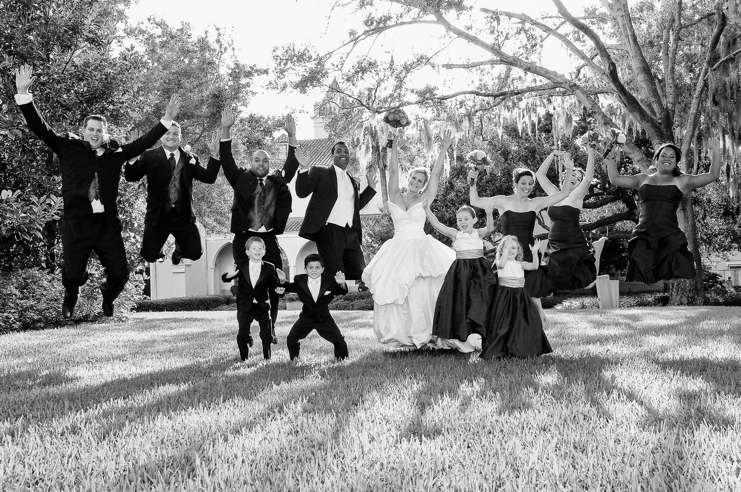 Wedding party jumping in celebration on grassy lawn in black and white.