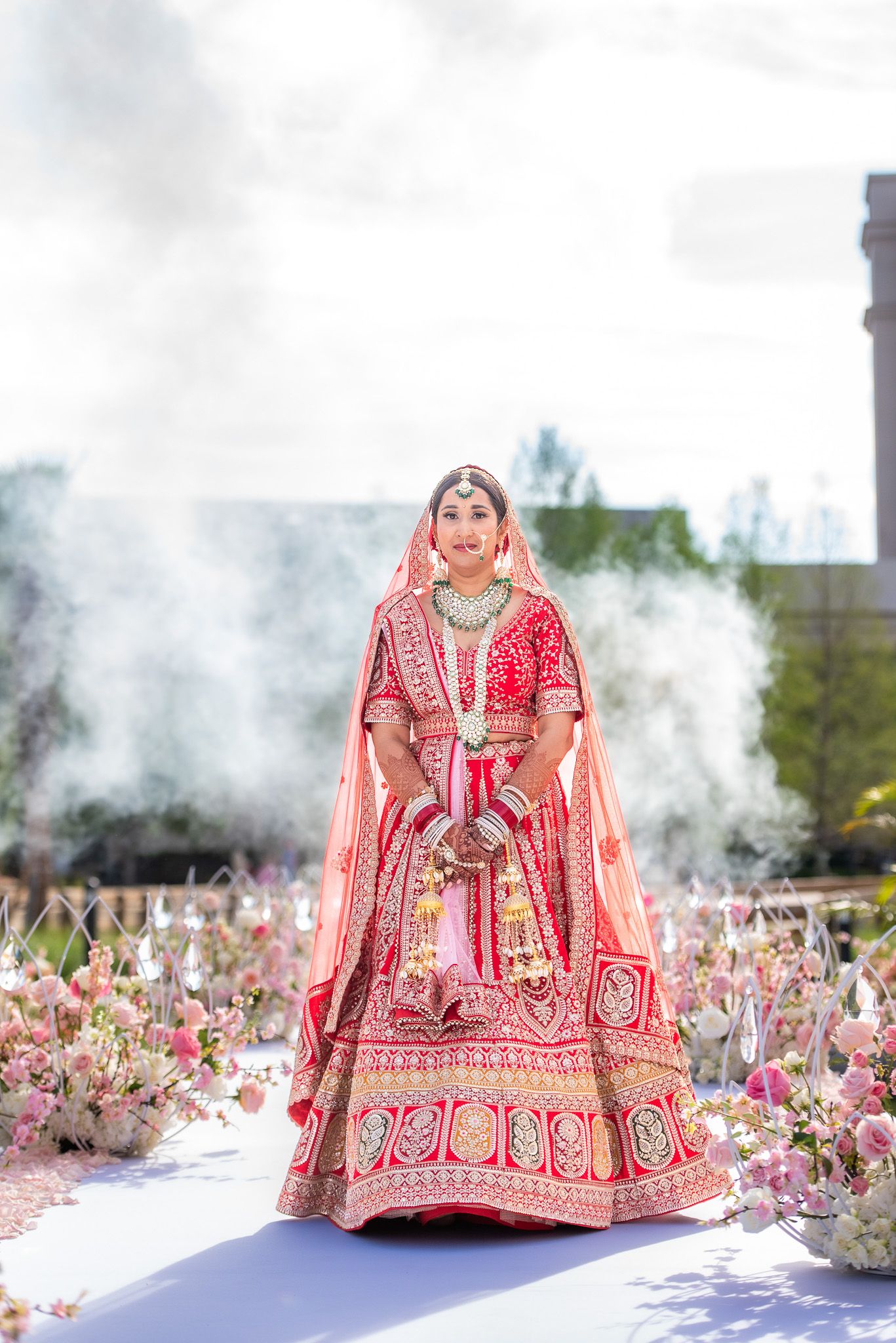 Bride in red lehenga poses amid smoke and floral decorations during outdoor wedding ceremony.