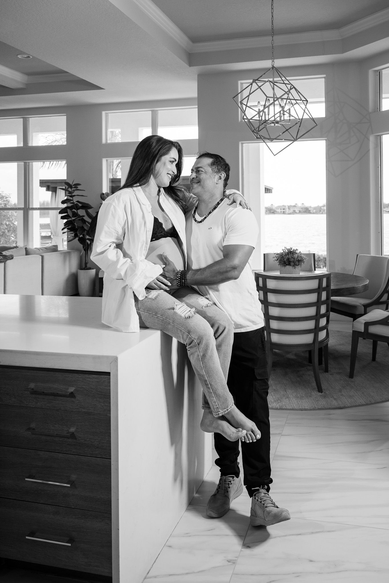 Intimate black and white photo of couple embracing in modern kitchen with large windows.