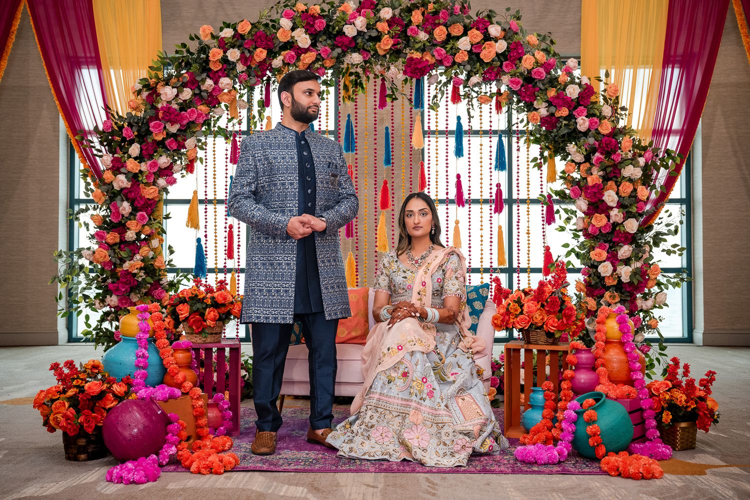 Traditional Indian wedding couple standing under ornate floral arch with bright decorations.