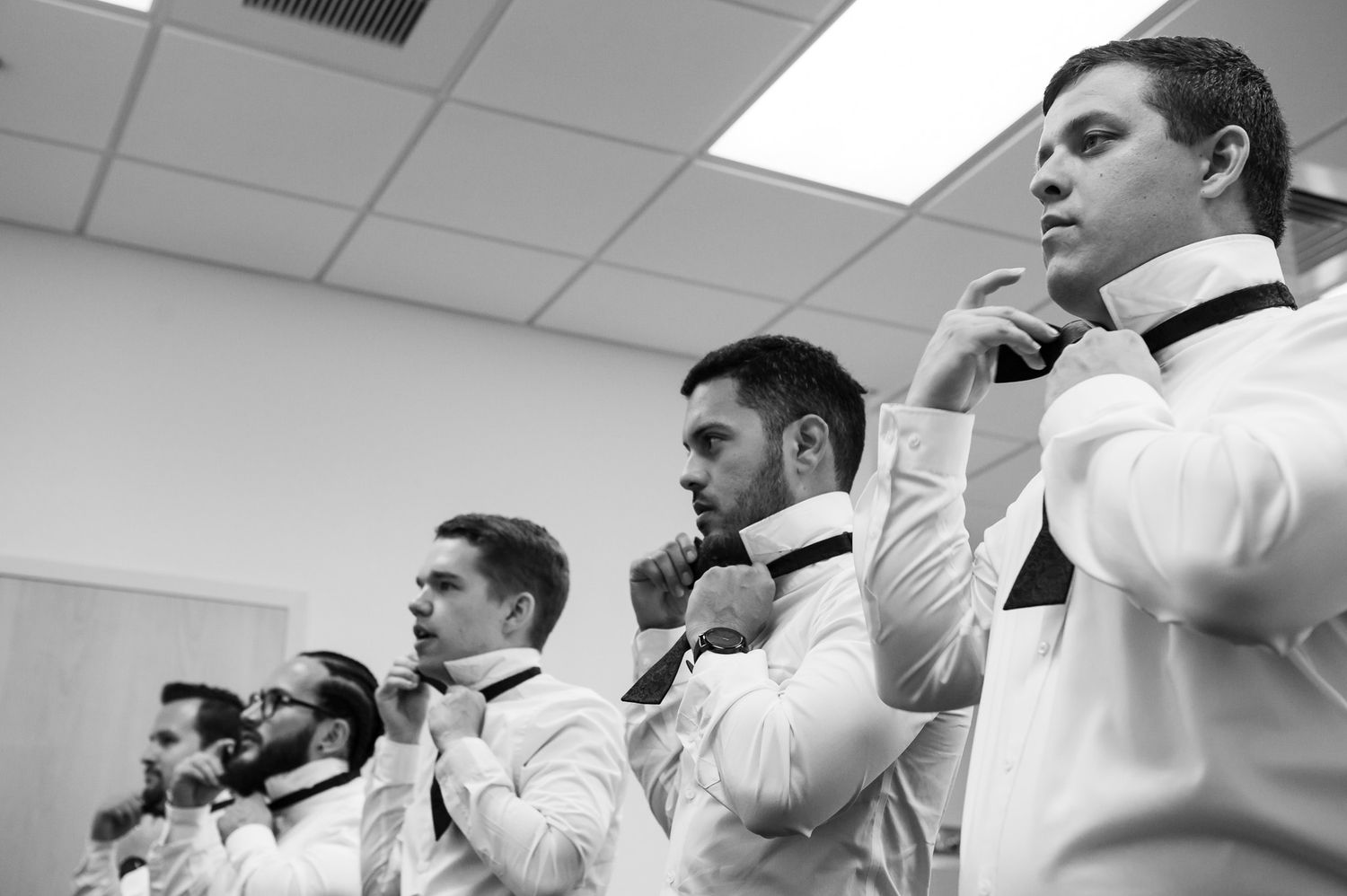 Row of groomsmen in formal white attire adjusting their ties before ceremony.