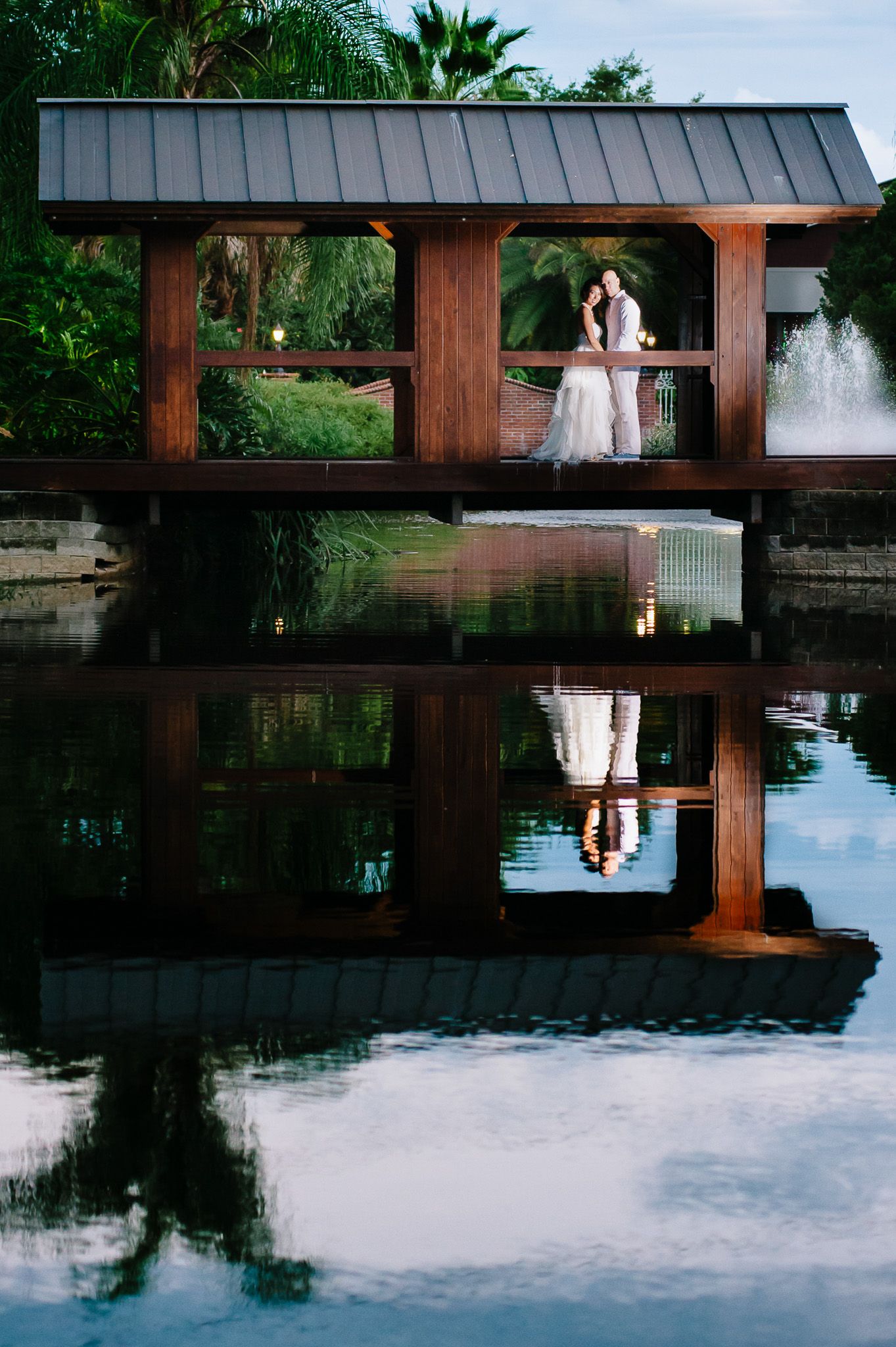 Wedding couple in covered bridge with reflection in pond below at tropical garden setting.
