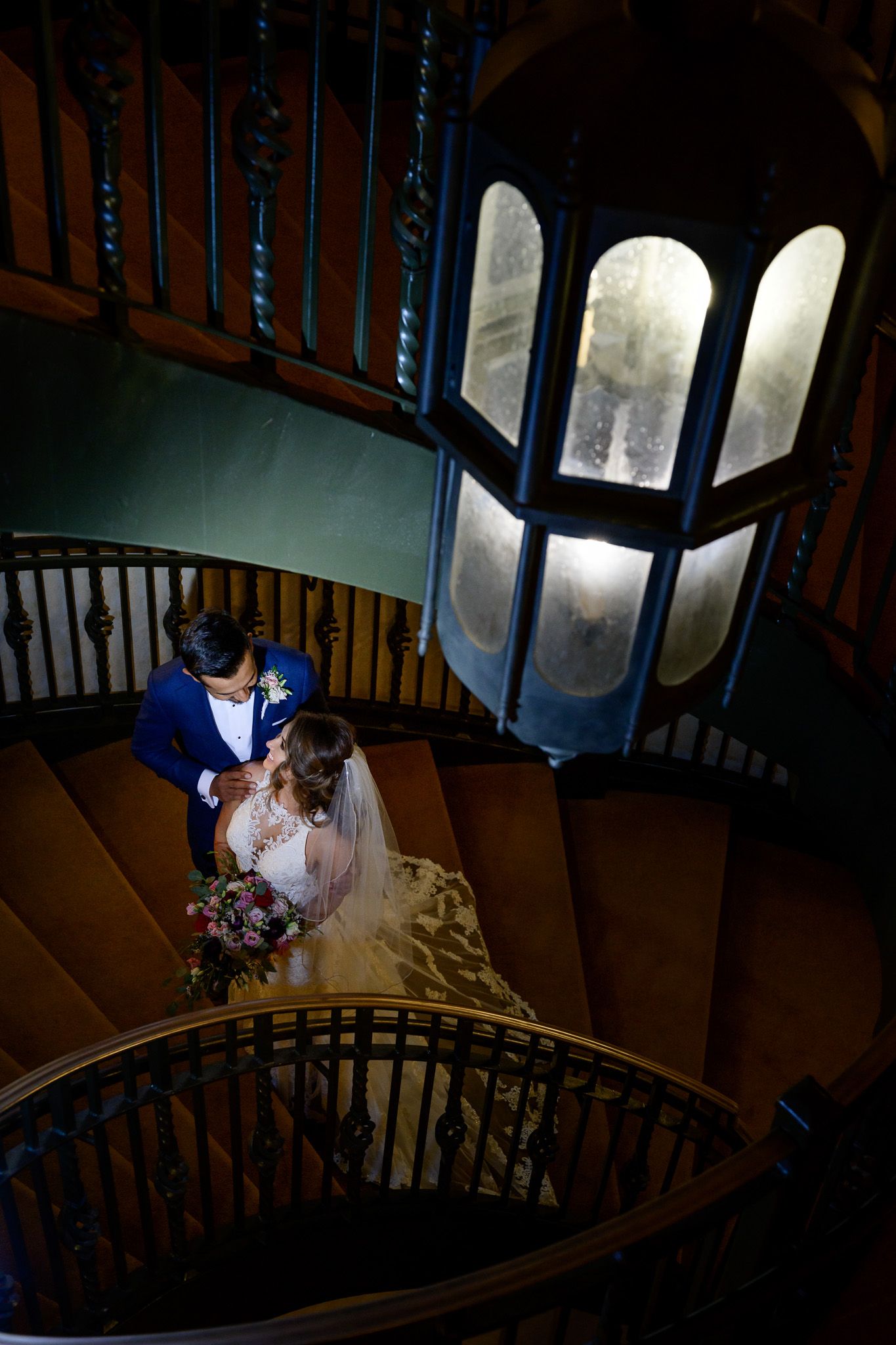 Dramatic wedding couple photo on spiral staircase with blue-lit window backdrop.