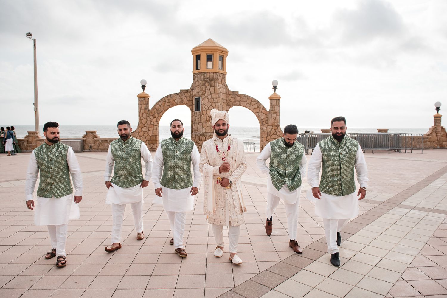Groomsmen in coordinating green and white outfits walking together under stone archway.