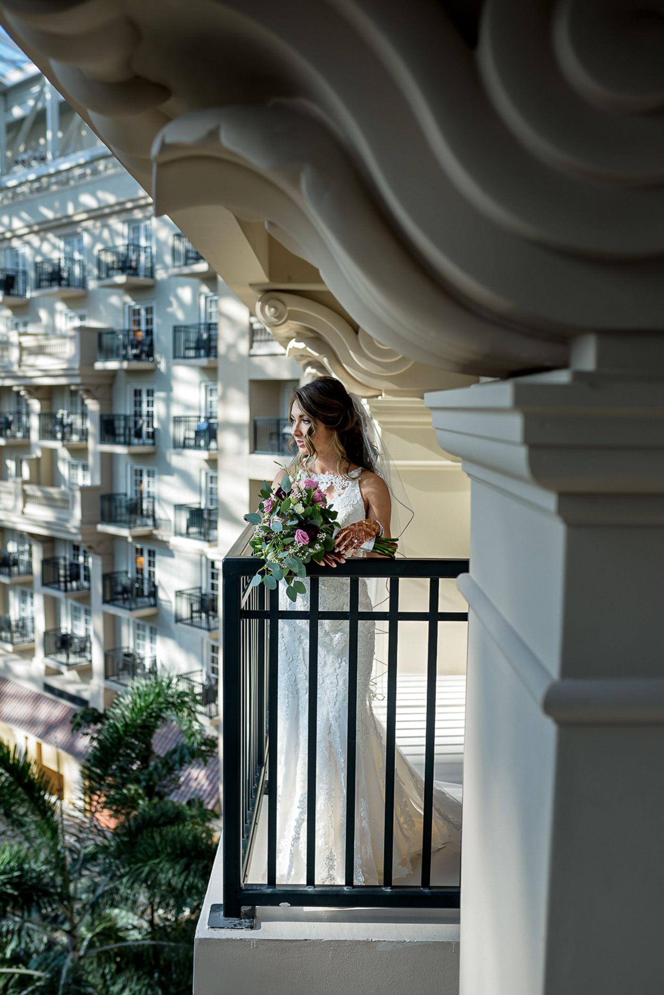 Bride holding bouquet on ornate balcony overlooking architectural details.