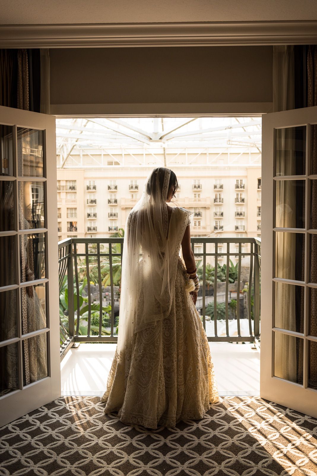 Silhouette of a bride in wedding gown standing at French doors overlooking a courtyard.