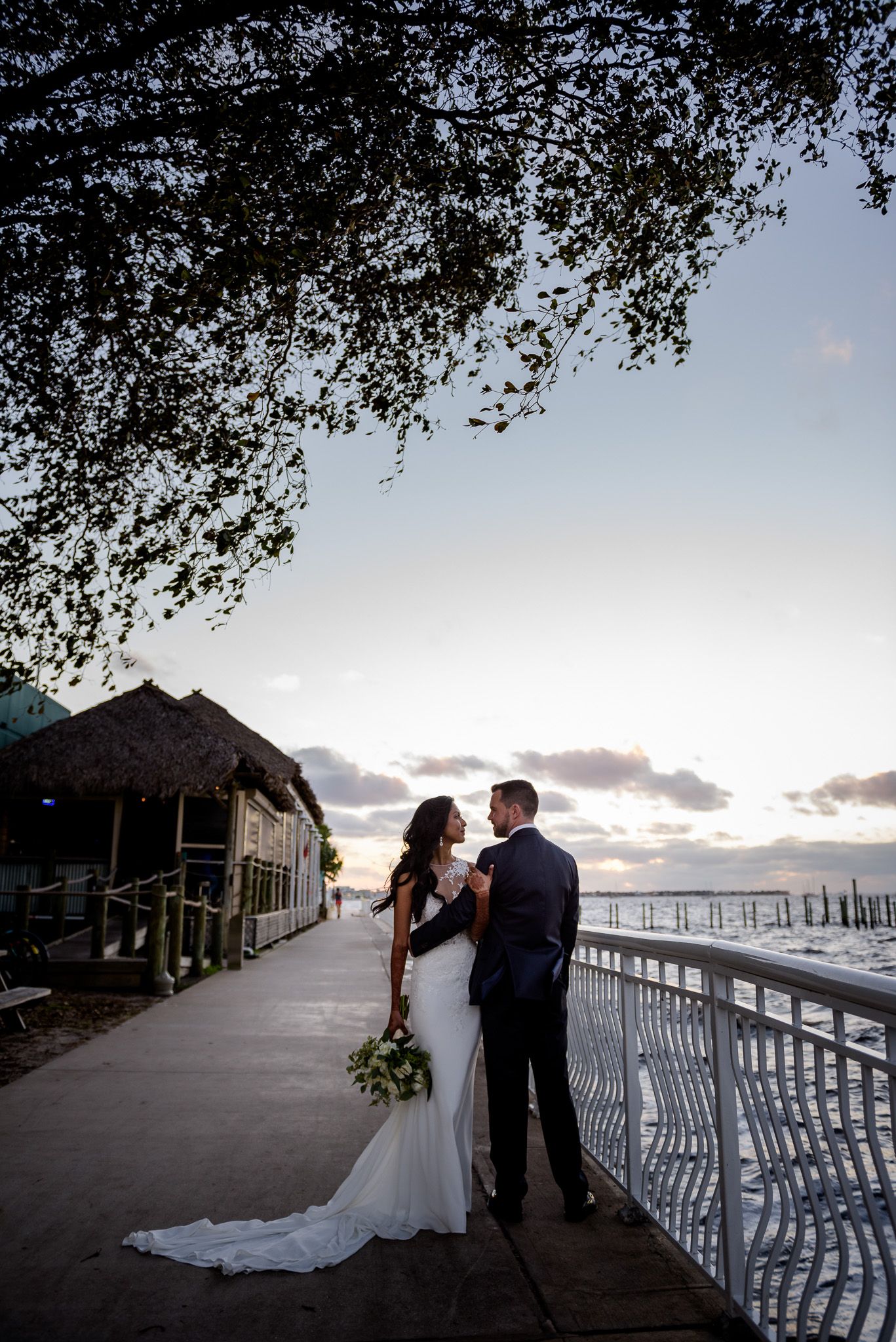 Newlyweds embrace on waterfront boardwalk at sunset with tree canopy overhead.