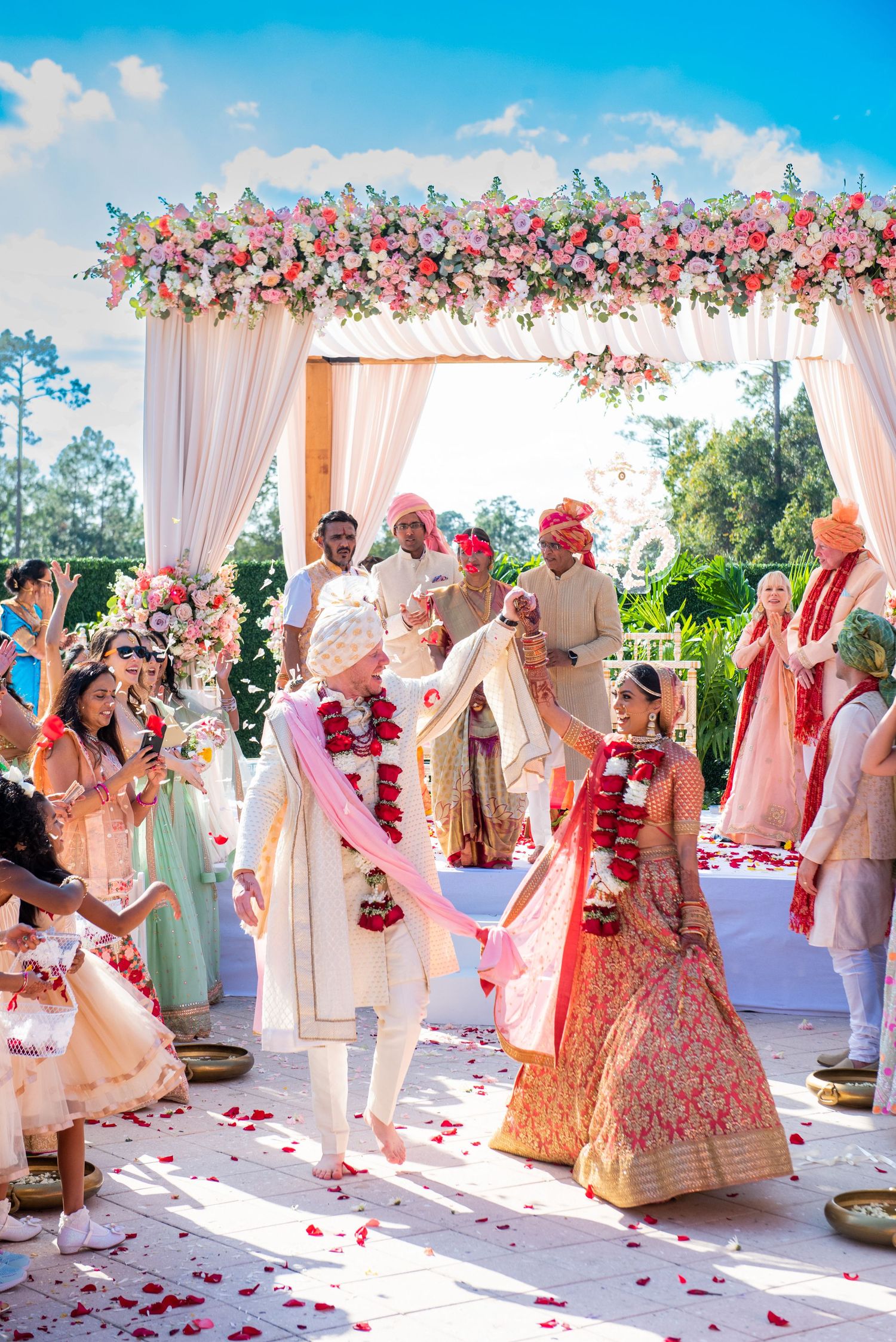 Outdoor wedding ceremony under floral mandap with pink and white draping and rose petals.