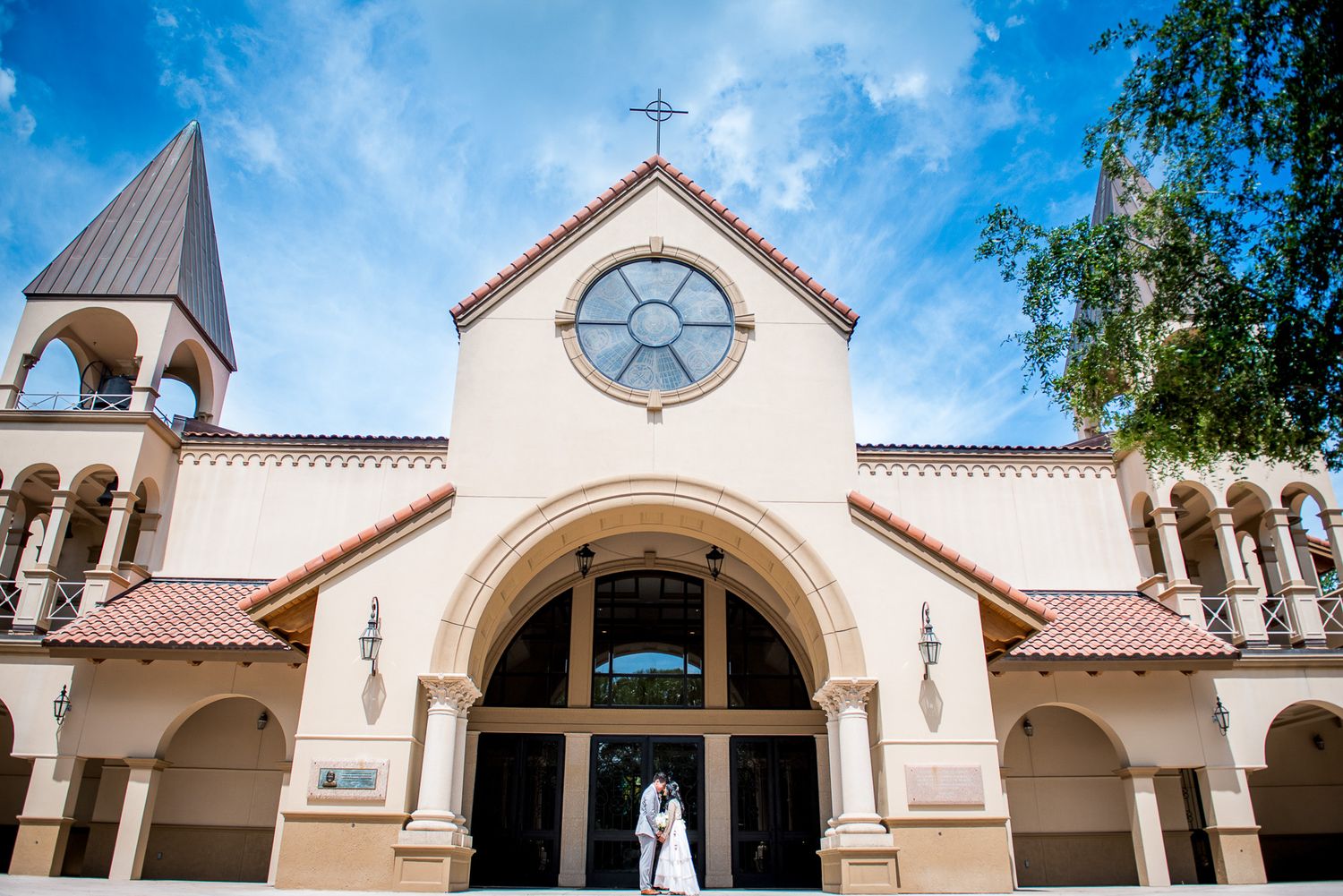 Wedding couple poses in front of Spanish-style church with bell tower under blue sky.