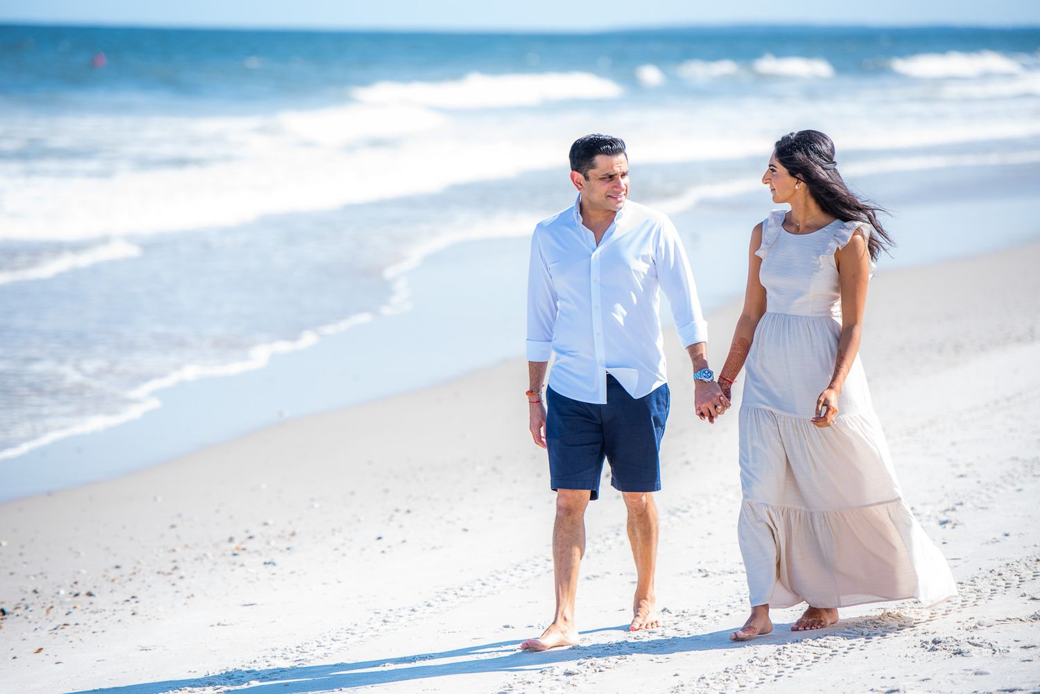 Casual beach engagement photo of couple walking along shoreline in light summer clothing.