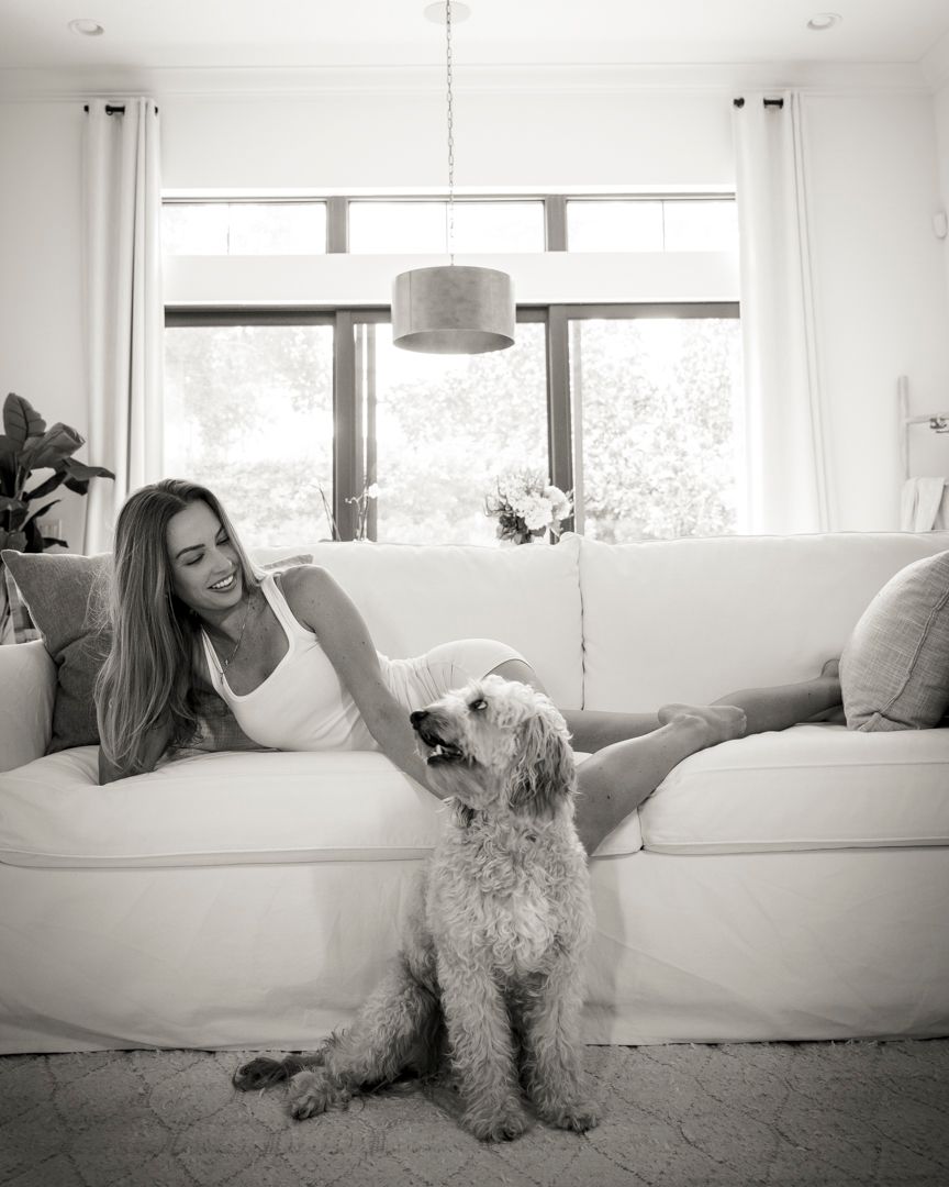 Woman relaxing on white couch with fluffy dog in bright living room.
