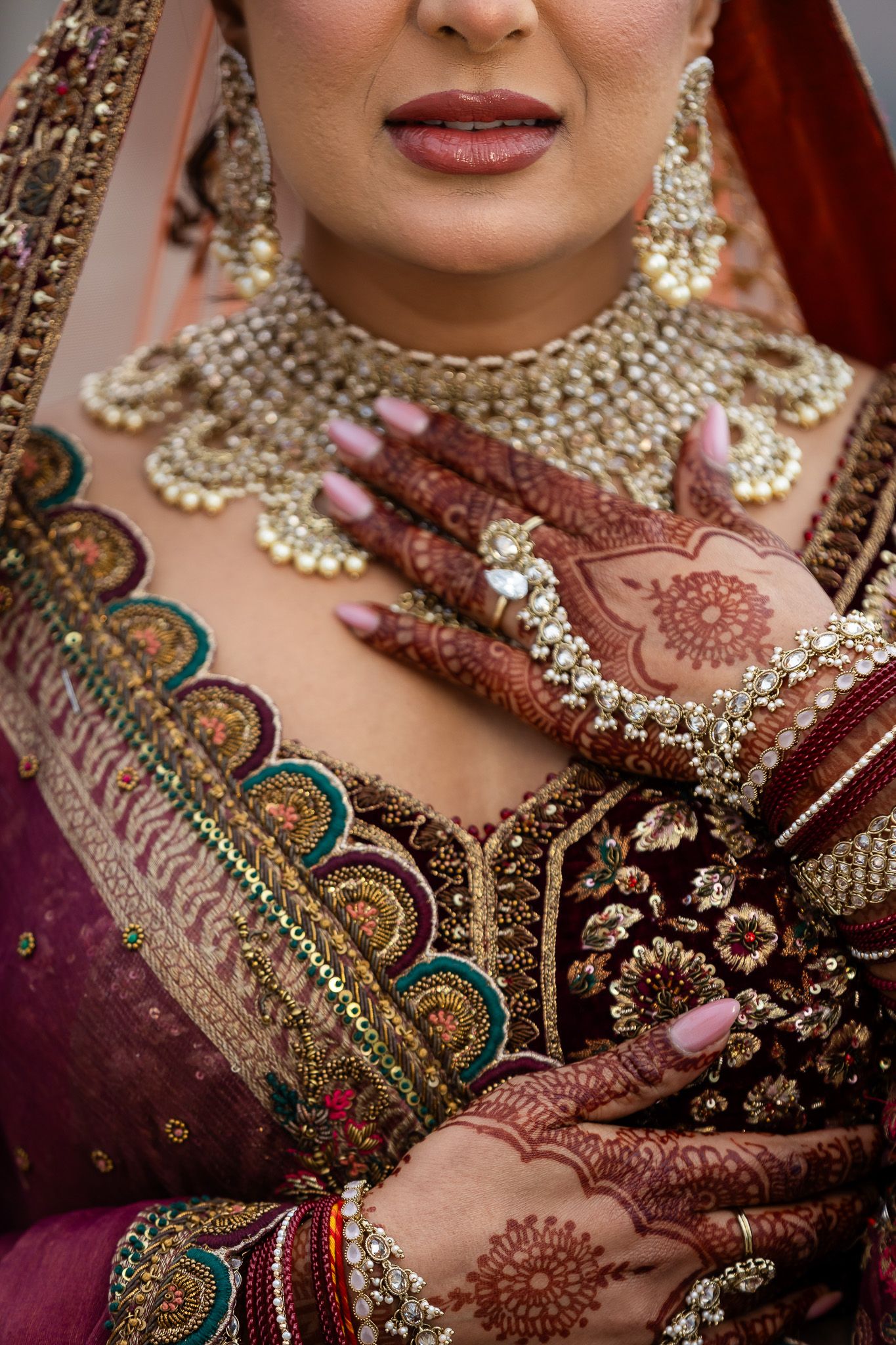 Close-up of Indian bridal jewelry and henna decorated hands with intricate traditional embroidered clothing.