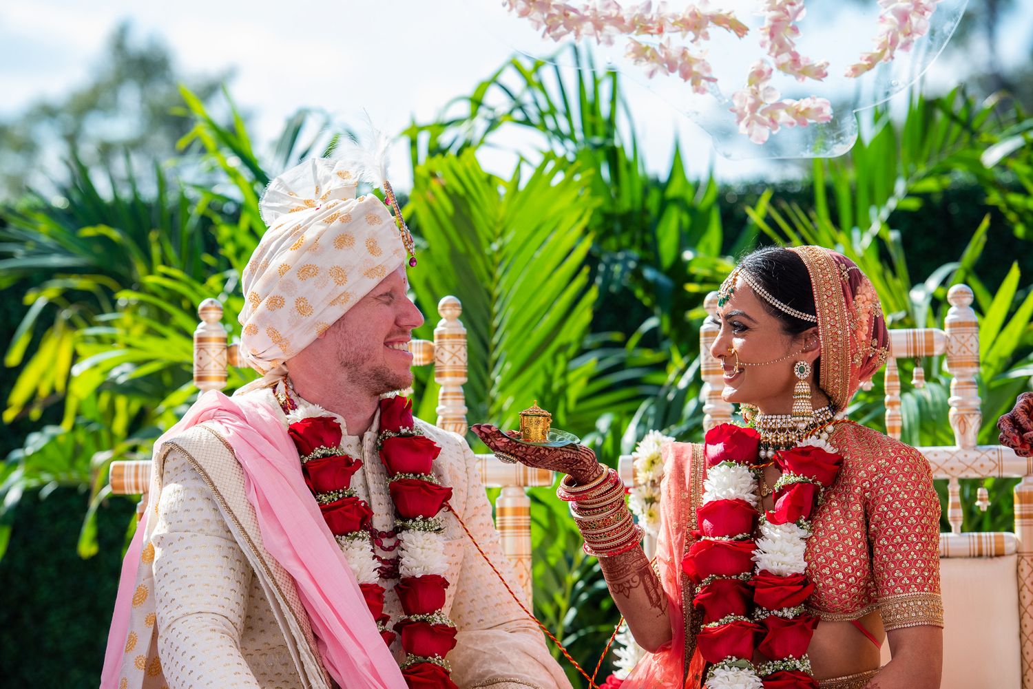 Traditional Indian wedding ceremony with red flower garlands amidst tropical palm leaves outdoors.