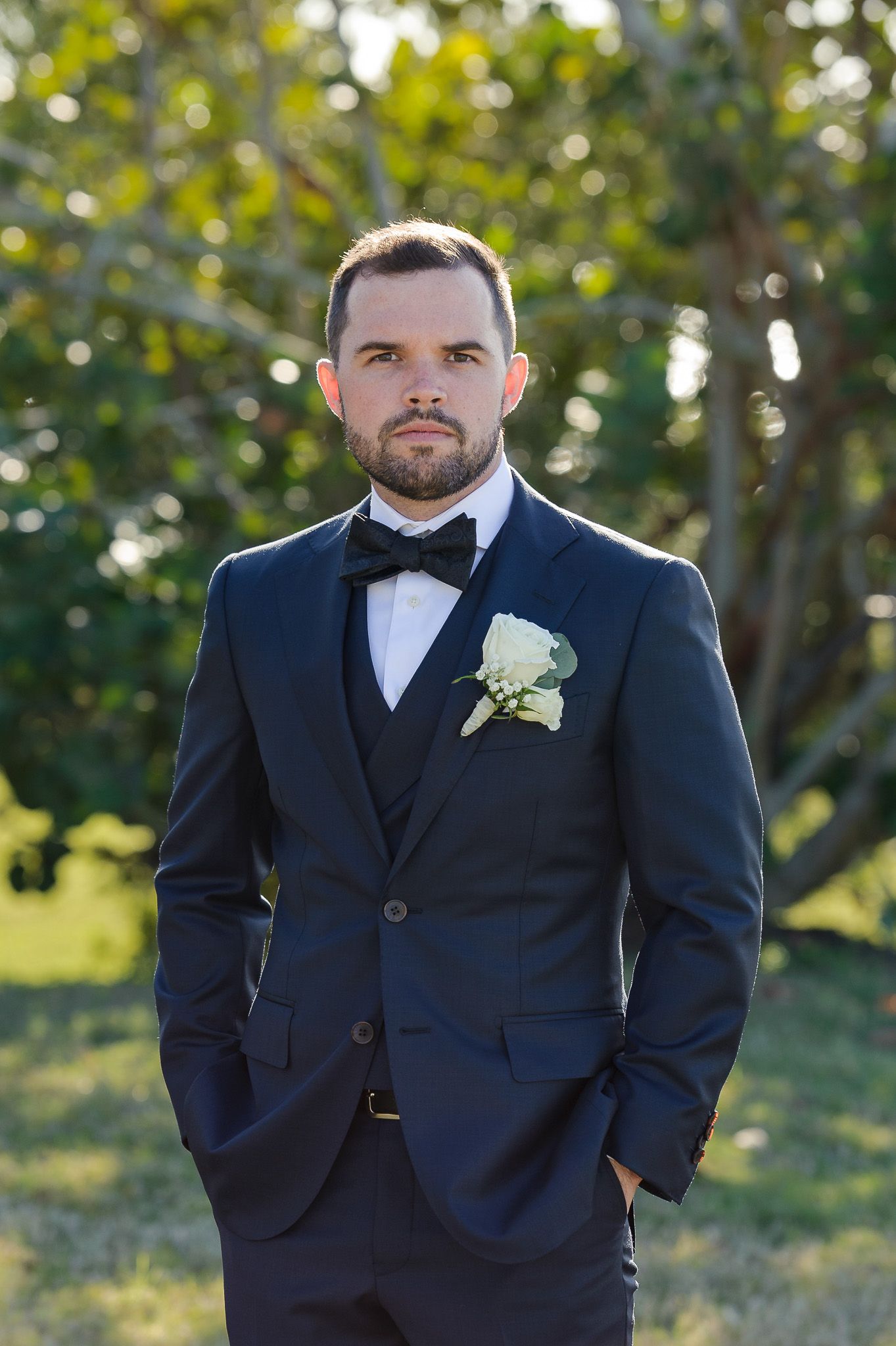 Formal portrait of person in navy tuxedo with white boutonniere against bokeh background.