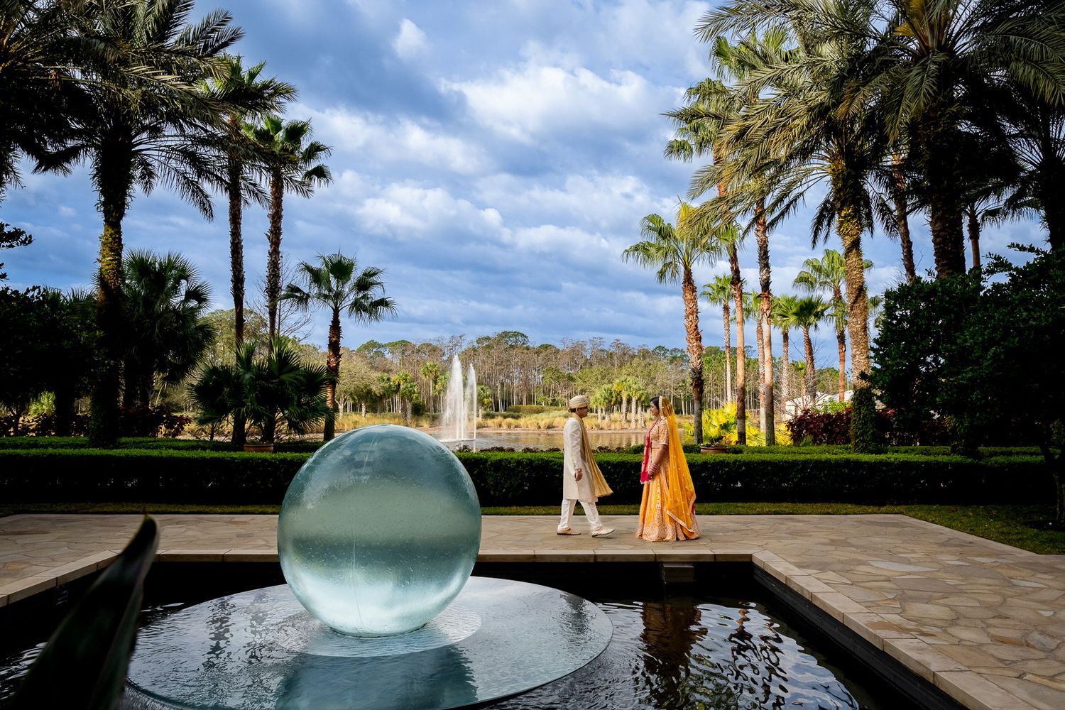 Modern glass sphere fountain with palm trees and blue sky in tropical wedding venue setting.