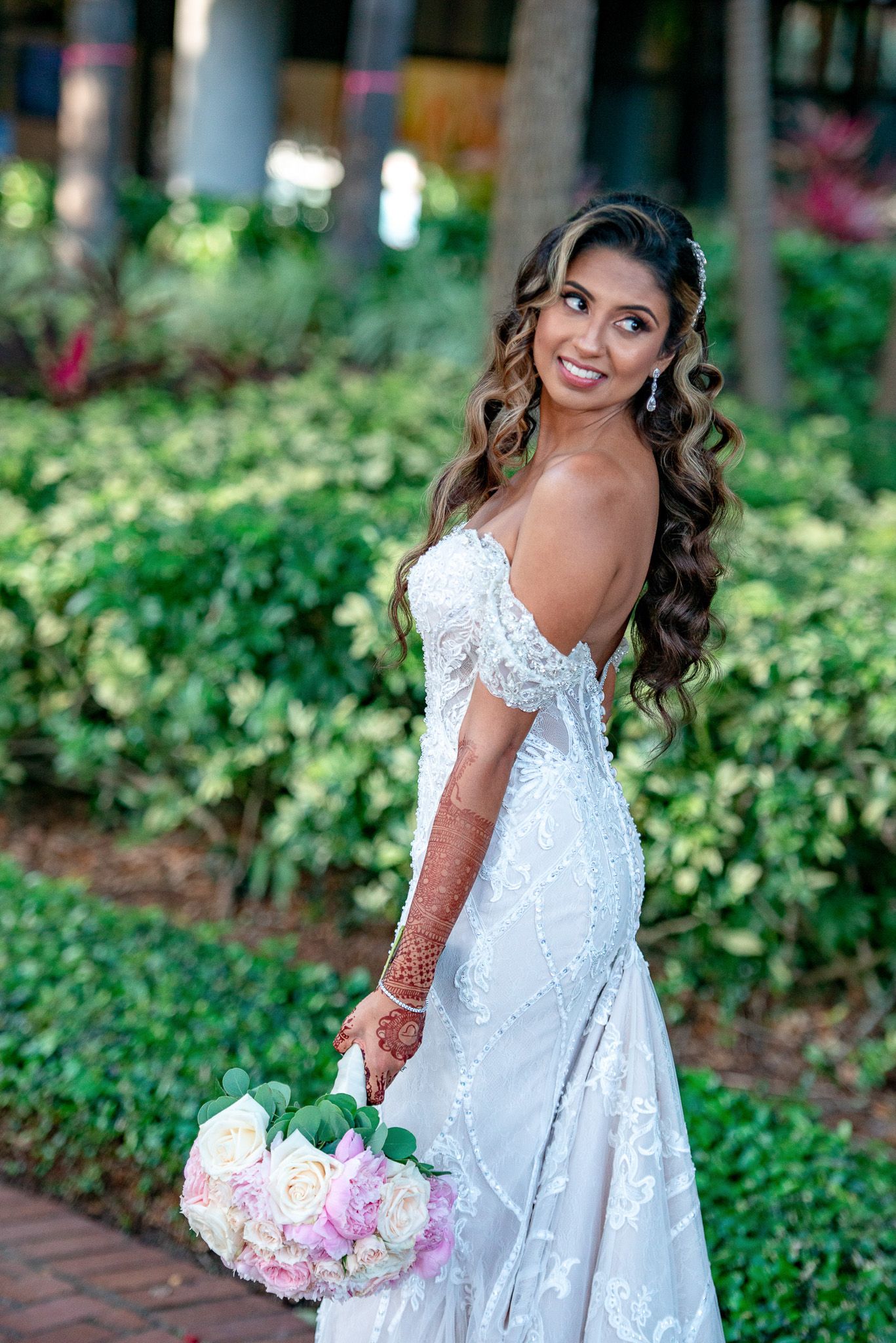 Bride in off-shoulder lace gown poses with bouquet against lush garden backdrop.