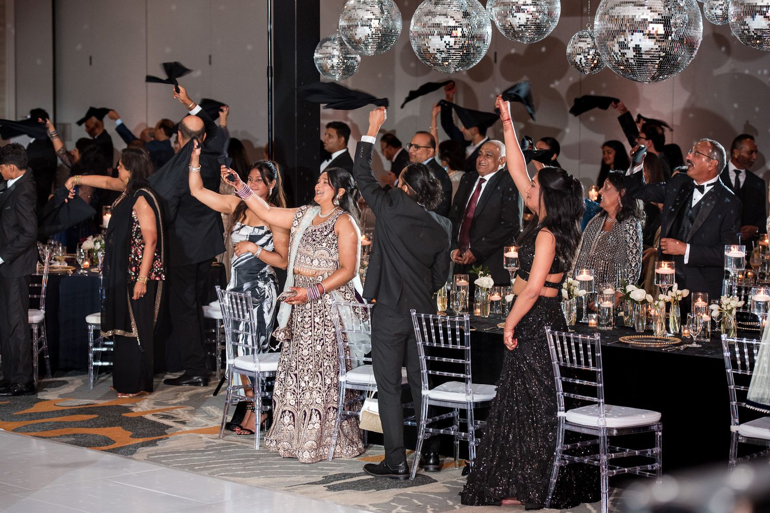 Large group celebrating in formal attire with raised hands under disco balls at an elegant event.