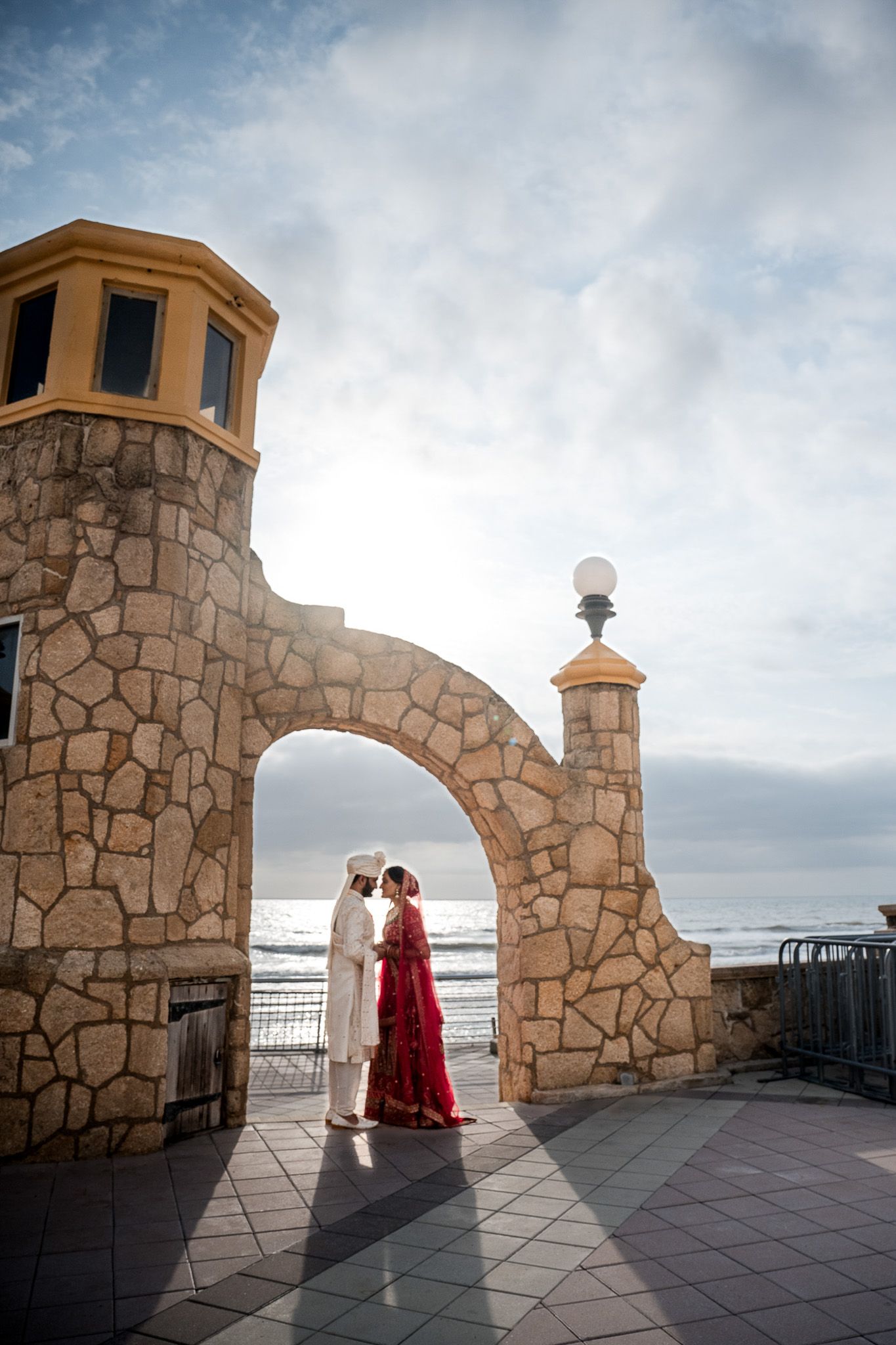 Wedding couple standing under stone archway against cloudy sky at scenic overlook.