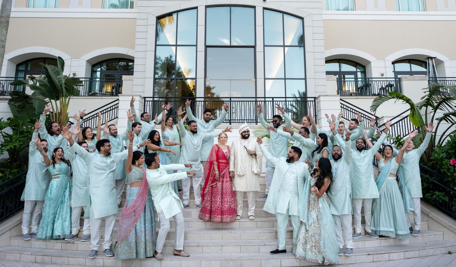 Joyful wedding party wearing mint green and white outfits celebrates on grand staircase with ornate architecture.