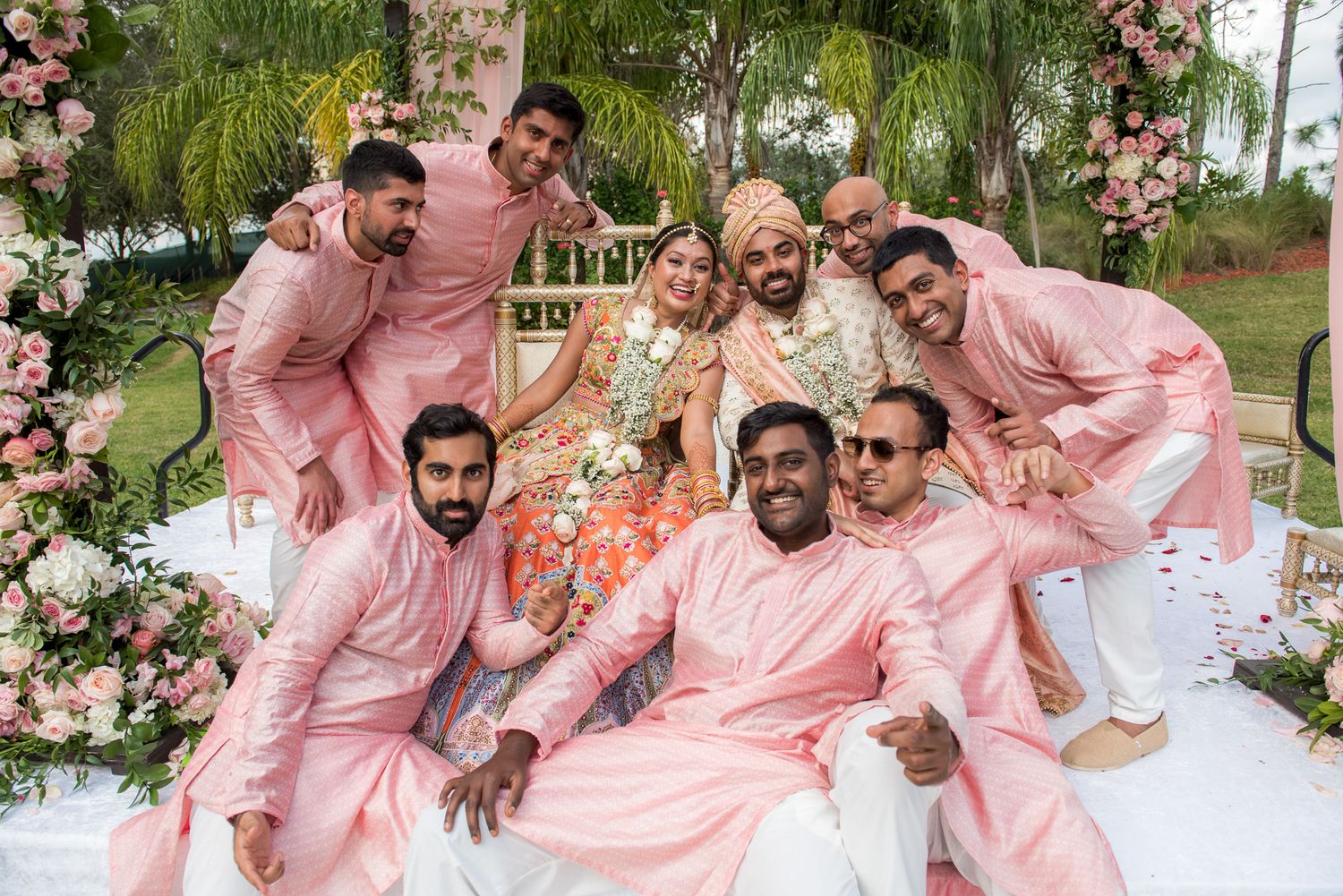 Wedding party in matching pink outfits poses together under floral arch.