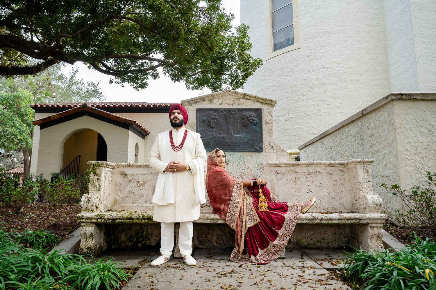 Traditional South Asian wedding couple posing on stone bench in garden setting.