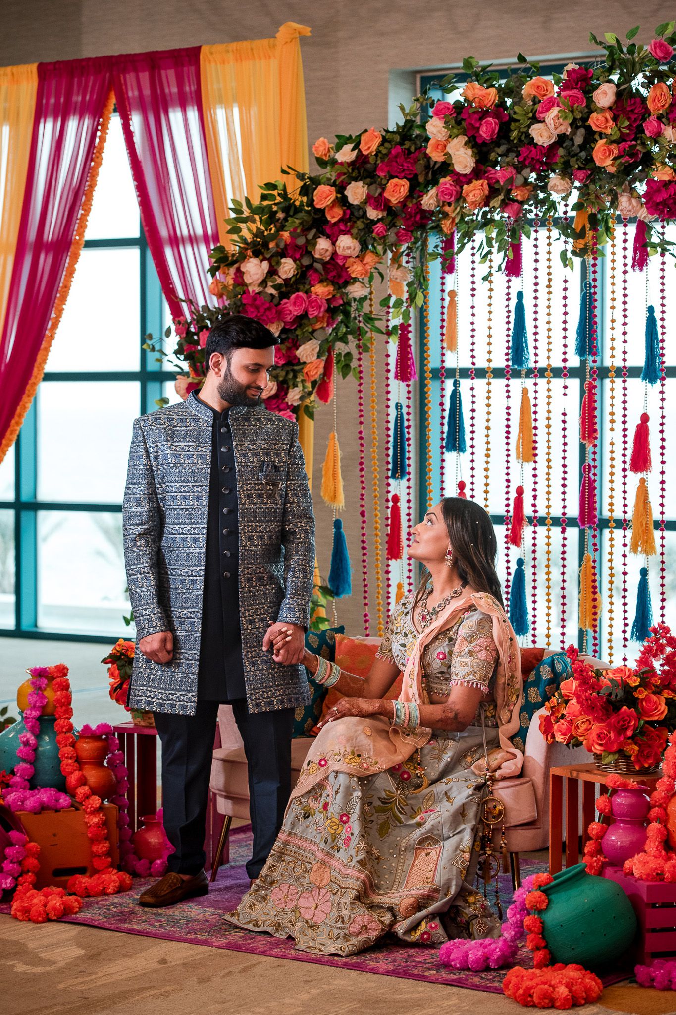Couple at Indian wedding ceremony under colorful floral arch with hanging tassels.