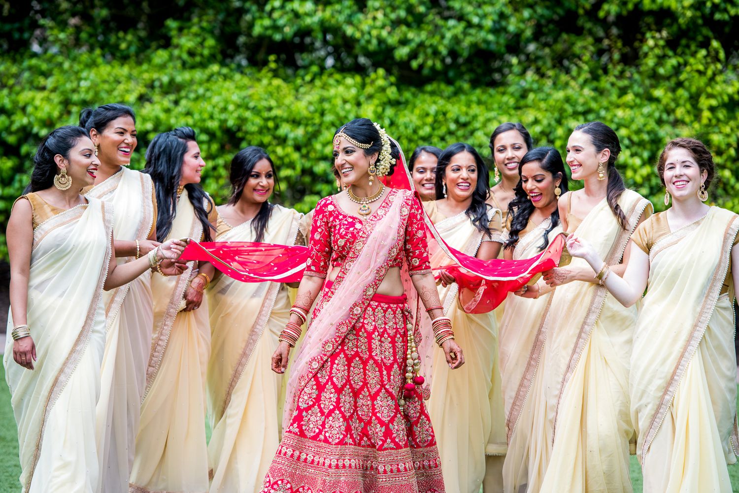Bride in red lehenga surrounded by bridesmaids in cream-colored sarees during outdoor photoshoot.