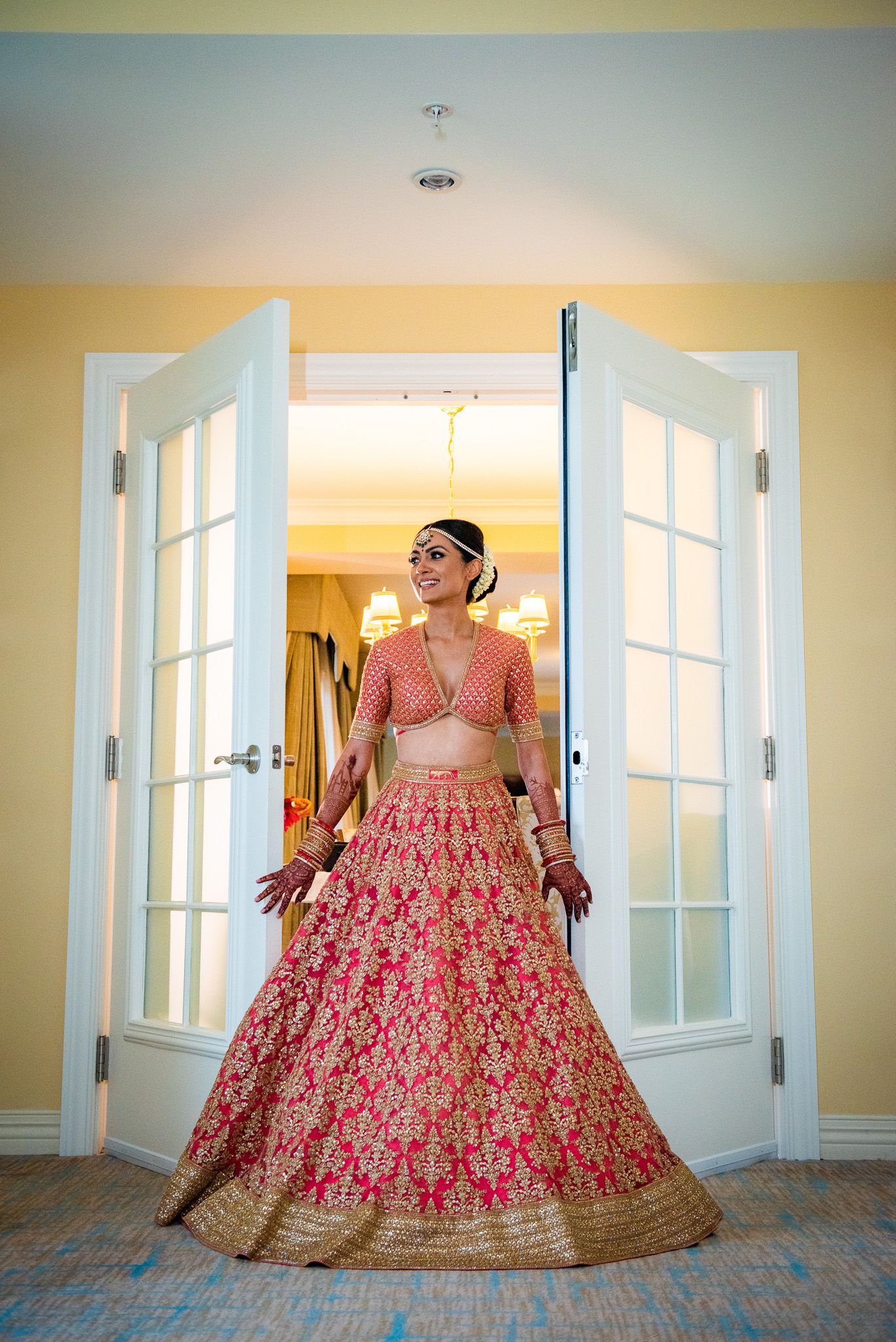 Bride wearing an ornate red and gold lehenga standing in front of white French doors.